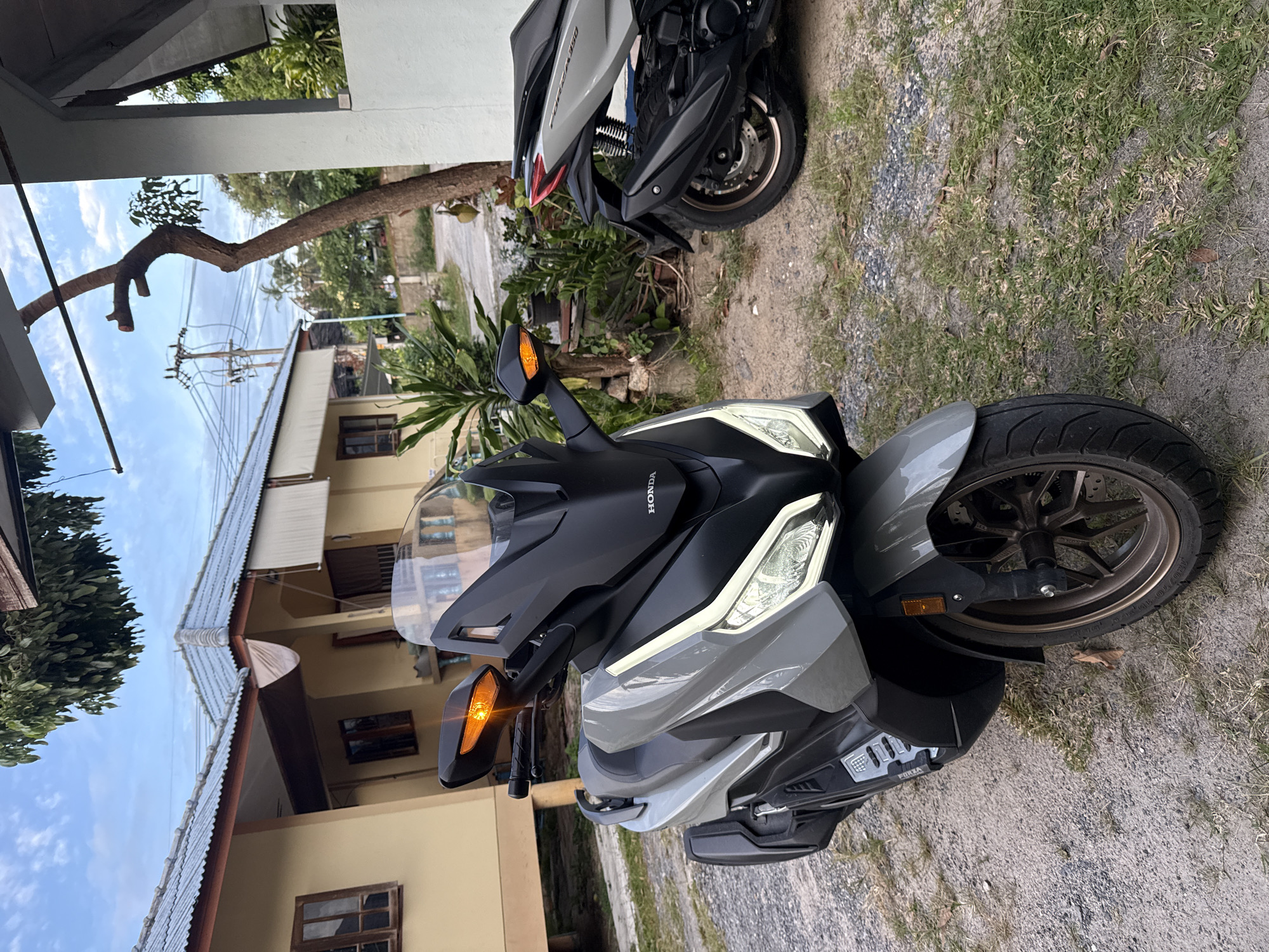 Two scooters are parked on a gravel and grass area in front of a light yellow house with a corrugated metal roof in the Ko Samui District, Thailand. The scooter in the foreground is gray and black with a large black windshield and LED headlights. The scooter in the background is silver and black. The sky is partly cloudy.