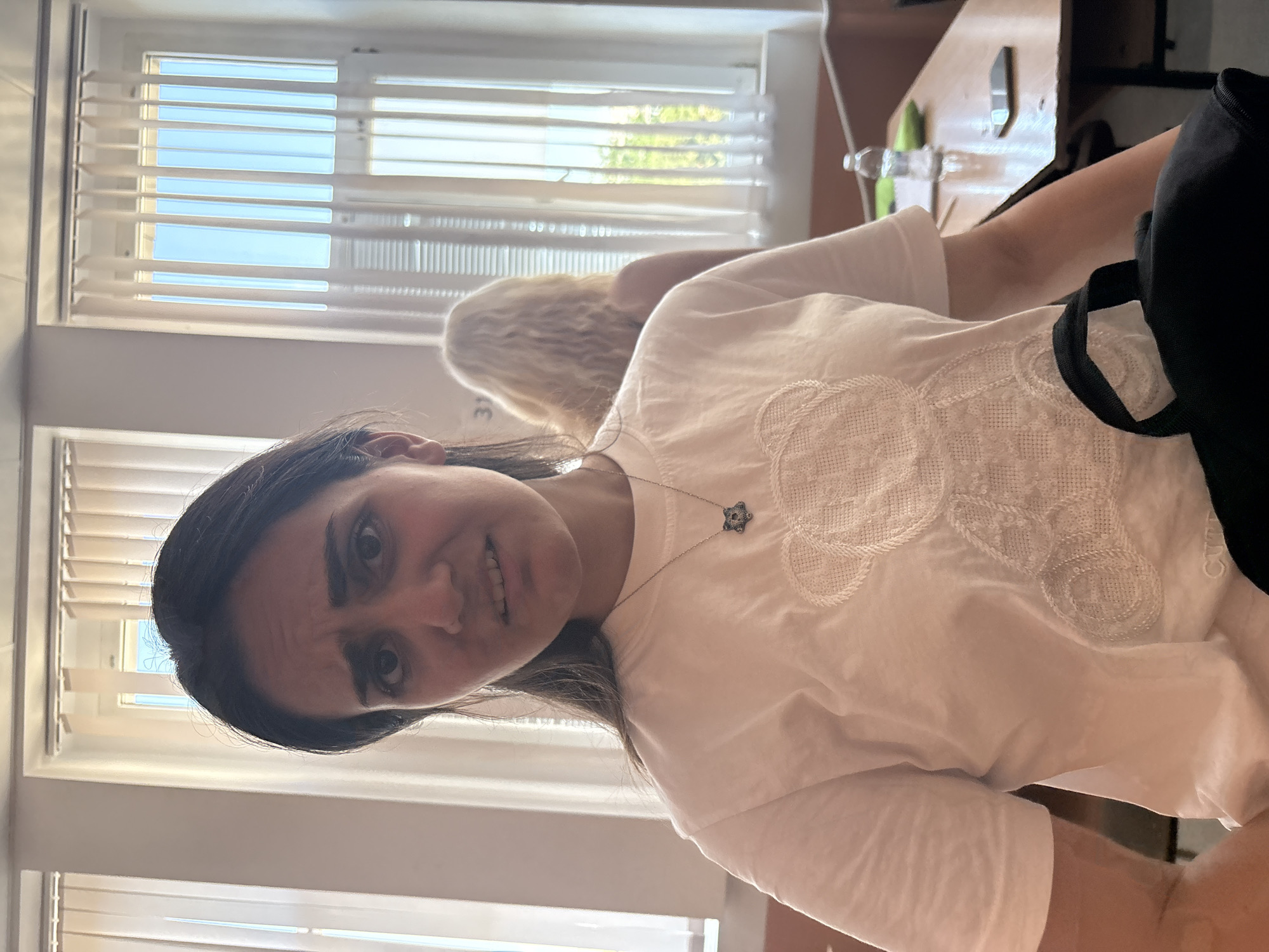 A woman with dark hair is wearing a white t-shirt with a decorative pattern and a necklace with a star-shaped pendant. She appears to be indoors. In the background are windows with blinds, and a second person with blonde hair is partially visible behind her. A desk or table is also visible. It is not possible to determine the specific location in Burgas, Bulgaria from the image.