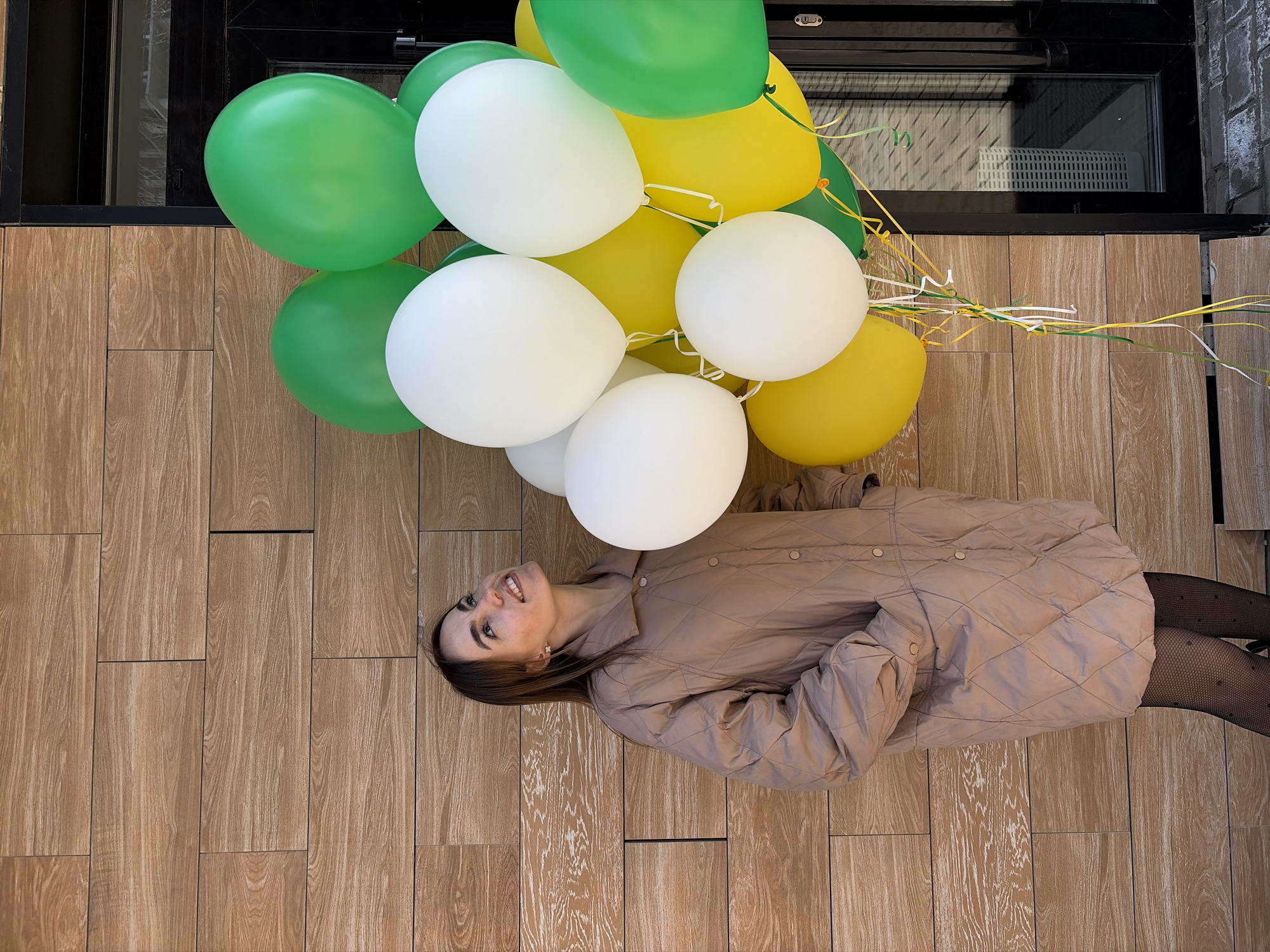 A woman in a tan quilted jacket and patterned tights lies on her back on a brown, wood-grained tiled surface. She holds a bunch of green, yellow, and white balloons. The location appears to be a building exterior in Izhevsk, Russia, judging by the urban architecture visible near the balloons and adjacent to the tiled surface.