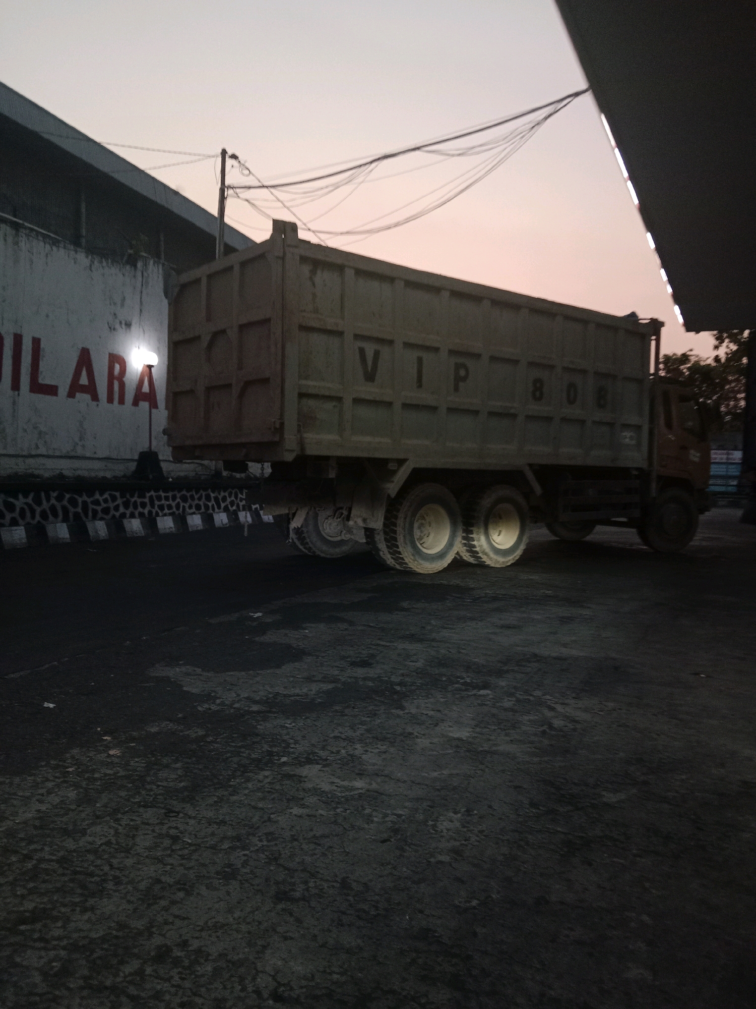 In Semarang, Indonesia, a parked dump truck with the markings "VIP 808" on its side is visible. The truck is a heavy-duty vehicle, likely used for construction or transport, and it is positioned on a dark, possibly paved, surface. Behind the truck, a textured concrete wall bears red lettering that appears to read "DILARA," suggesting a prohibition or warning. Overhead, a network of power lines crisscrosses the sky, indicating an urban or industrial setting. The ambient light suggests it is either dawn or dusk, with a pale, soft sky. The scene is devoid of human presence, focusing solely on the static presence of the truck and its surroundings. The overall atmosphere is quiet and still.