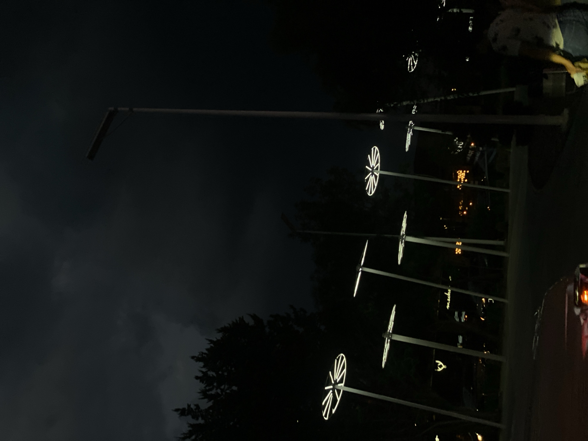 At night in Unknown, Unknown, a row of illuminated, wheel-shaped lights line the road, with a standard street lamp at the head of the row. A car is visible in the lower right corner. Dense trees and a dark, cloudy sky fill the background. A person stands near the head of the line of lights.