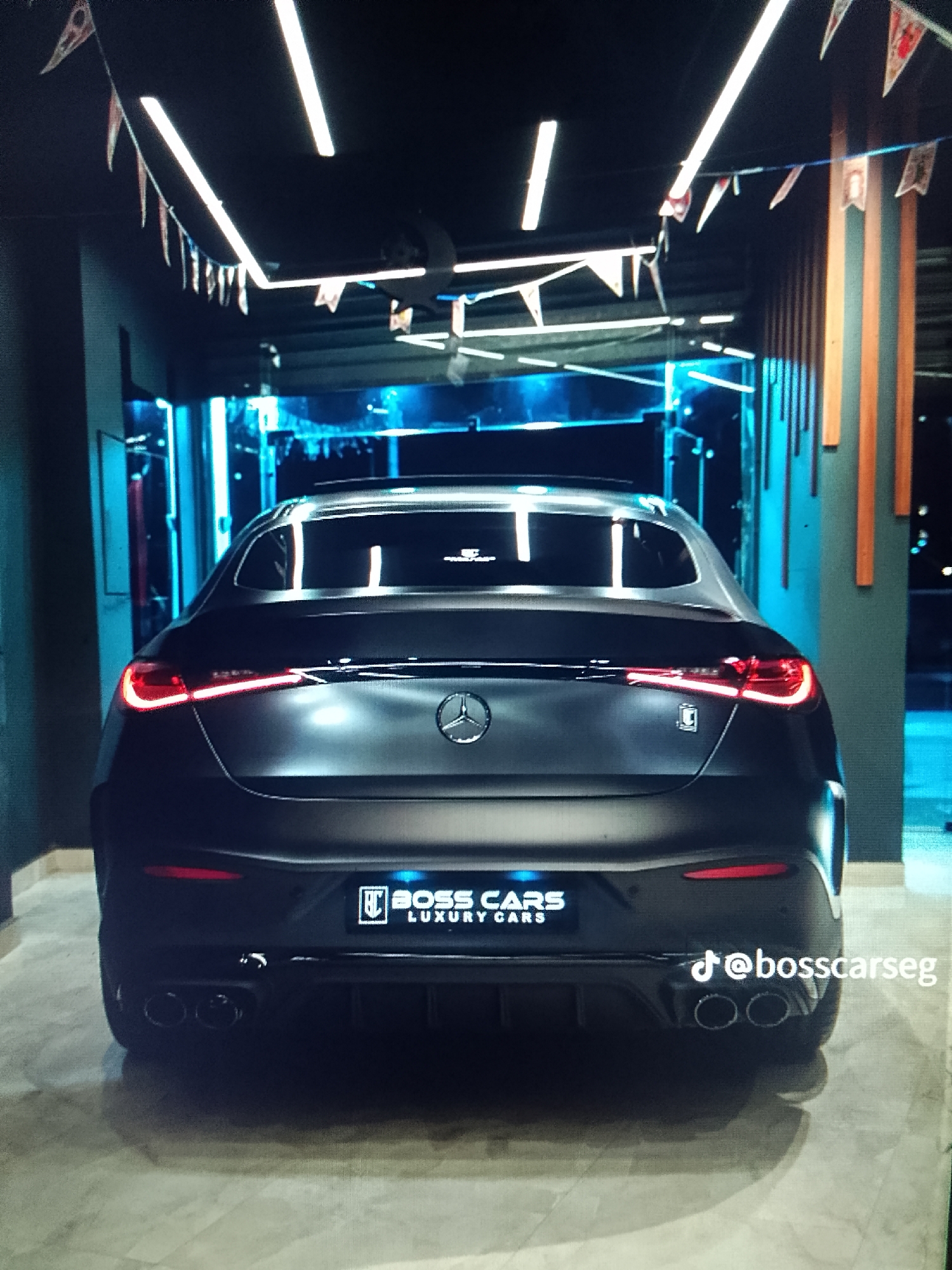The image depicts the rear of a matte black Mercedes-Benz luxury vehicle parked indoors in Unknown, Unknown. The vehicle's red taillights are illuminated, and a prominent Mercedes-Benz emblem is centered on the trunk. Below the emblem, a custom license plate area displays "BOSS CARS LUXURY CARS" with a stylized "BC" logo. A small, square badge with a stylized "B" or logo is affixed to the right side of the trunk near the taillight. The vehicle features a rear diffuser and quad exhaust tips. Above the vehicle, multiple bright, linear LED lights are mounted on a dark ceiling. Pennant flags, primarily white and red, are strung across the ceiling as decorative elements. The background features blue accent lighting, possibly from vertical LED strips, and structured walls with alternating vertical panels of a light wood-like material and a darker material on the right. The floor is a light-colored, reflective surface. No individuals are visible. A TikTok handle, "@bosscarseg," is overlaid in white text on the lower right of the image.