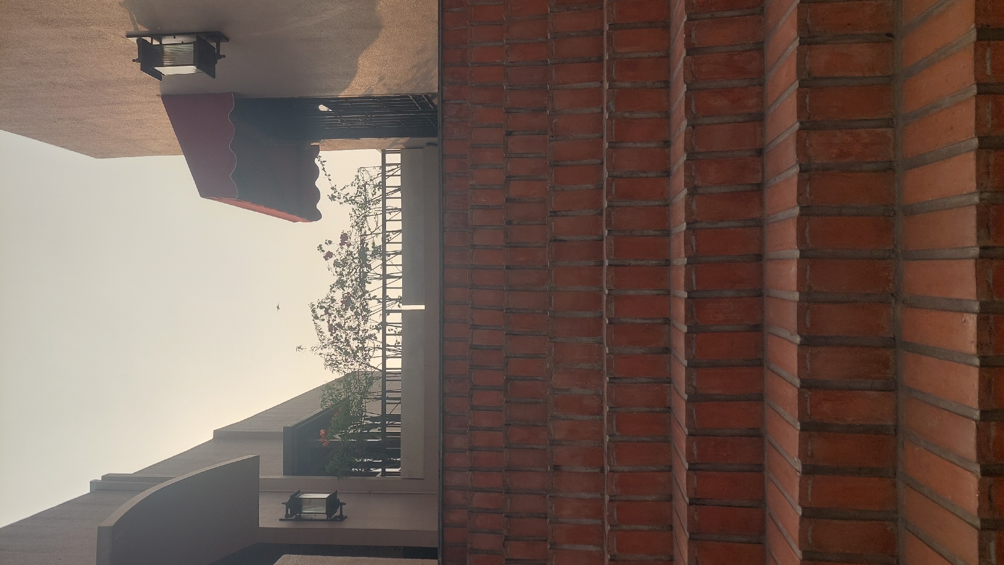The image depicts an outdoor scene featuring a wide, multi-tiered staircase made of red bricks with gray mortar. The perspective is looking up the stairs towards a balcony or upper level. Above the stairs, there is a building with a tan-colored facade. To the left, a portion of a building with an arched overhang is visible, with a wall-mounted lantern. A red awning is attached to the tan building on the right, above a dark, barred doorway. Lush green plants with small red flowers are visible on the balcony behind a railing. The sky is a pale, hazy yellow, suggesting either early morning or late afternoon light, or possibly atmospheric conditions. A small bird is in flight in the upper center of the image. The scene appears to be a quiet, architectural exterior, possibly part of a residential complex or a commercial establishment in Cileungsi, Indonesia.