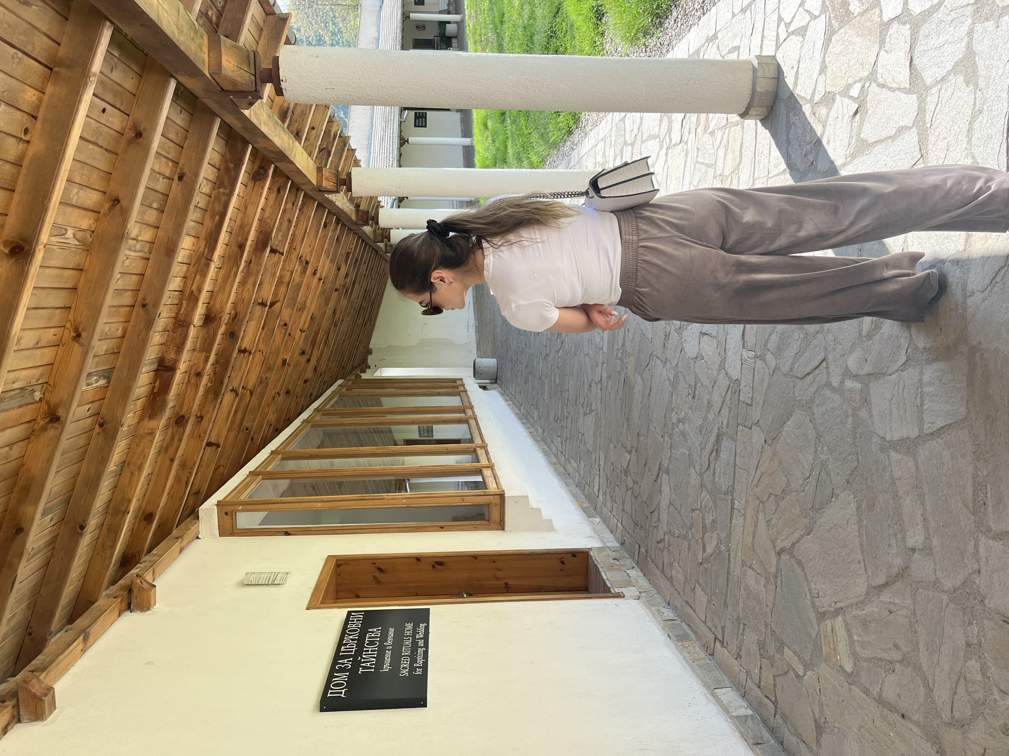 A full shot depicts a woman standing on a stone-paved walkway next to a building in an unidentified location in Bulgaria. The walkway consists of irregularly shaped grey stones. The woman is angled to the left, wearing sunglasses, a white t-shirt, and brown sweatpants. She also has a white purse with a chain strap slung over her shoulder.
To the left of the walkway is a building. The building is white with a wooden roof and a row of windows near the roof. Below the windows, there is a sign with black lettering on a black background that reads "ДОМ ЗА ЦРКОВНИ ТАИНСТВА" in Cyrillic, and "SACRED RITUALS HOME for Baptism and Wedding" in English.
Along the edge of the walkway are large, white, cylindrical columns. Beyond the columns, there is a grassy area and another building in the background. The overall setting suggests a historical or cultural site.