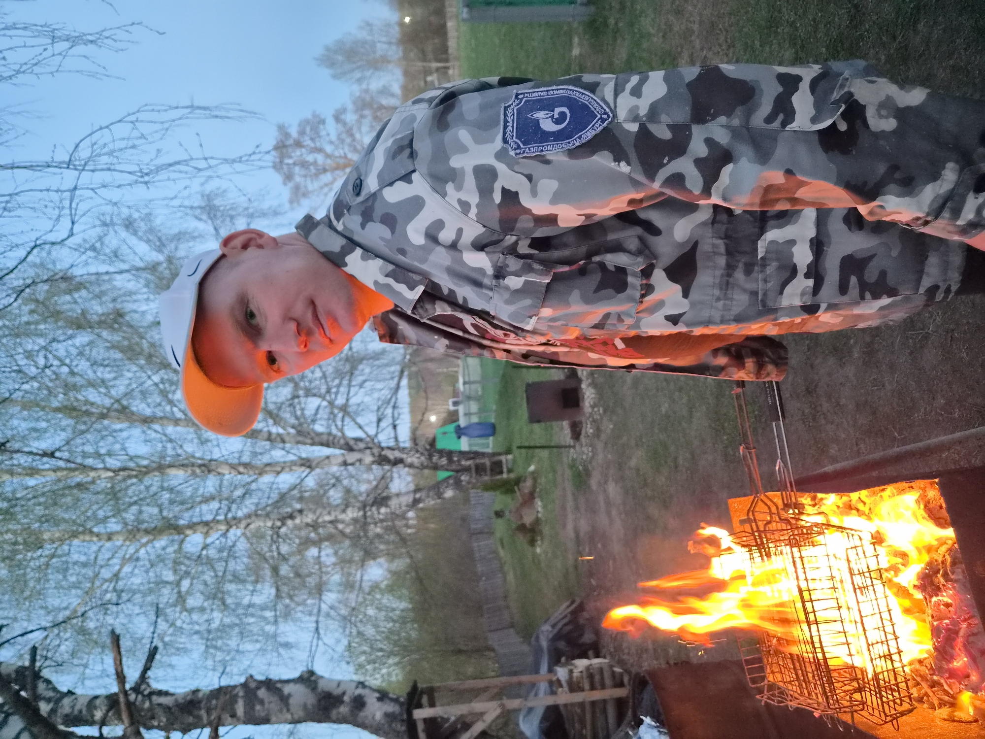 The image depicts a man in a camouflage jacket and a white baseball cap standing next to a barbecue grill with flames. The man is looking towards the camera. The jacket has a patch on the sleeve. The location appears to be outdoors, possibly in a rural or suburban setting, with trees and a fence visible in the background. Based on information in the patch, the location is Узяр, Russia.