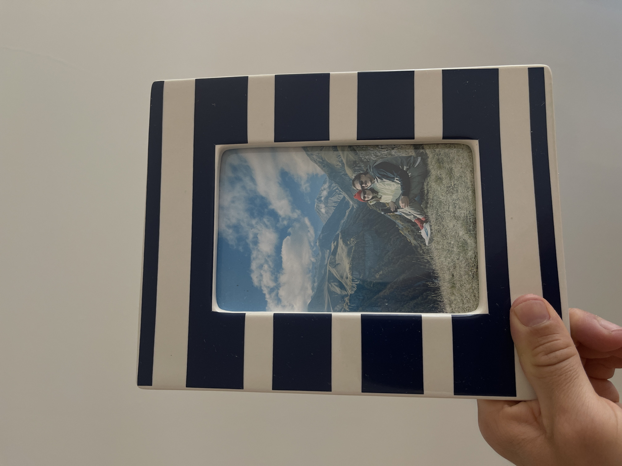 A person holds a framed photograph. The frame is striped with alternating navy blue and white stripes. The photograph inside shows a couple in a mountainous, possibly alpine, landscape with visible sky, mountains and sparse vegetation. The couple is wearing outerwear; one person has on a red hat and red shoes. The background is a plain white surface. The image does not contain any information that identifies the city of Malinovka, Russia.