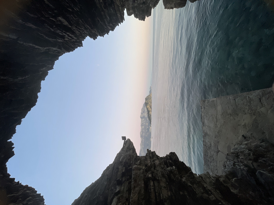 The image captures a coastal landscape framed by a natural rock archway or cave opening along the Budva Riviera in Budva, Montenegro, with a clear view of Sveti Nikola Island.
The scene is viewed from within a dark rock formation, which frames the upper, left, and right portions of the image. These rock formations exhibit distinct horizontal stratification and irregular, jagged edges. On the lower left, a light-colored, flat, rectangular concrete or stone platform extends into the clear, blue-green seawater. A small, dark, cubic object protrudes from the left rock face.
The midground is occupied by the Adriatic Sea, which appears calm with minor surface ripples. The water near the foreground platform is translucent, revealing submerged features.
The background presents a clear sky transitioning in color from light blue at the zenith to a faint pinkish-purple near the horizon, indicative of twilight. A thin, white linear mark, consistent with an aircraft contrail, is visible in the upper right quadrant of the sky.
In the distance, Sveti Nikola Island, characterized by its green vegetation and rocky profile, is prominently visible. To the island's left, a mountainous coastline is discernible, featuring what appears to be coastal settlements and a small white boat on the water. Additional mountains extend along the distant horizon behind the island. No human figures or discernible activities are observed.