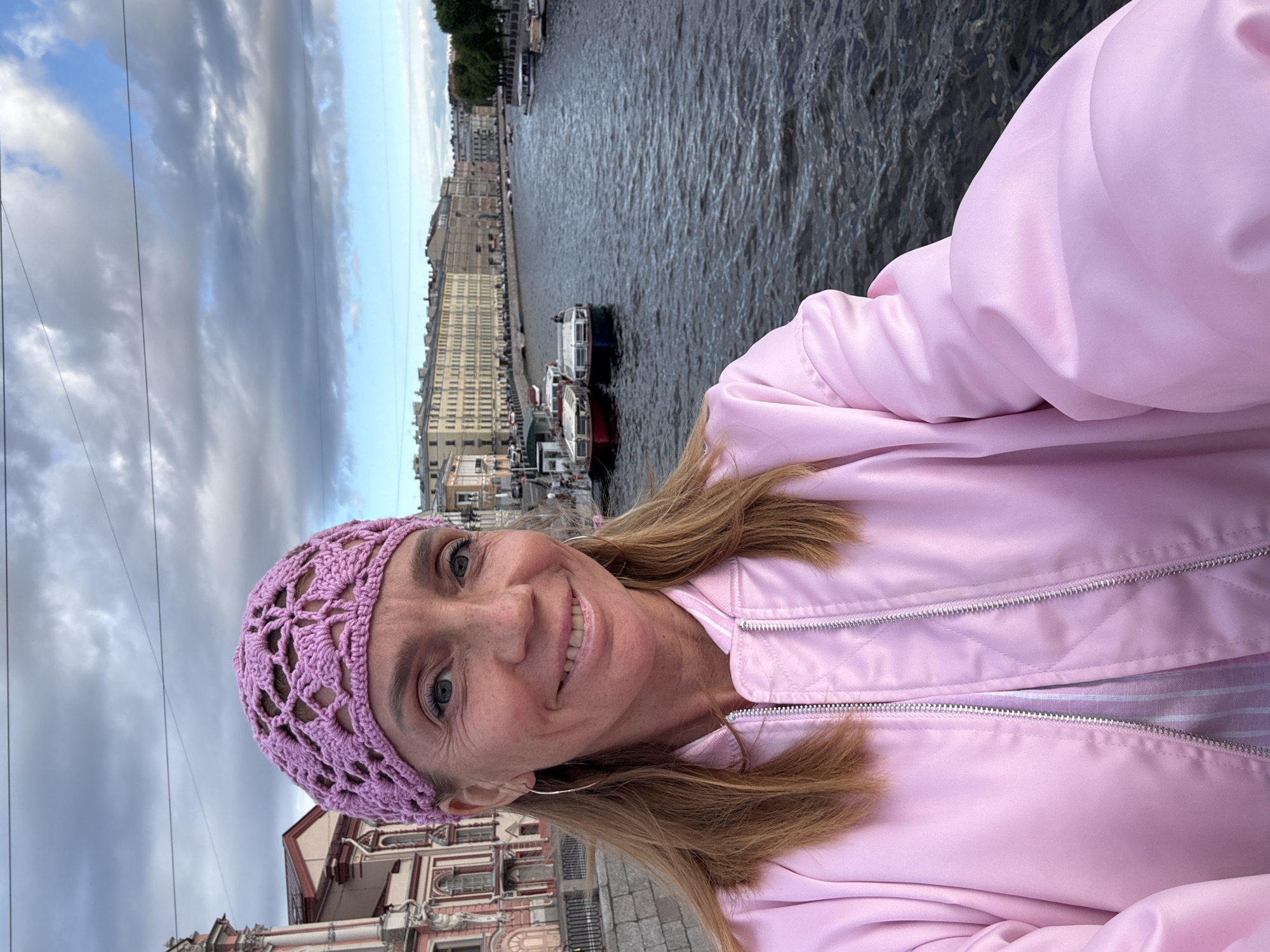 A woman is taking a selfie on a bridge in Saint Petersburg, Russia. She is wearing a pink knitted hat and a pink satin bomber jacket. The woman has blonde hair and blue eyes. She is smiling and looking at the camera. In the background, there is a canal with boats and buildings on either side. The sky is cloudy. The canal is the Griboyedov Canal. The buildings are part of the historic architecture of Saint Petersburg. The boats are typical of those used for sightseeing tours on the canals. The cloudy sky suggests that the weather is cool and overcast.