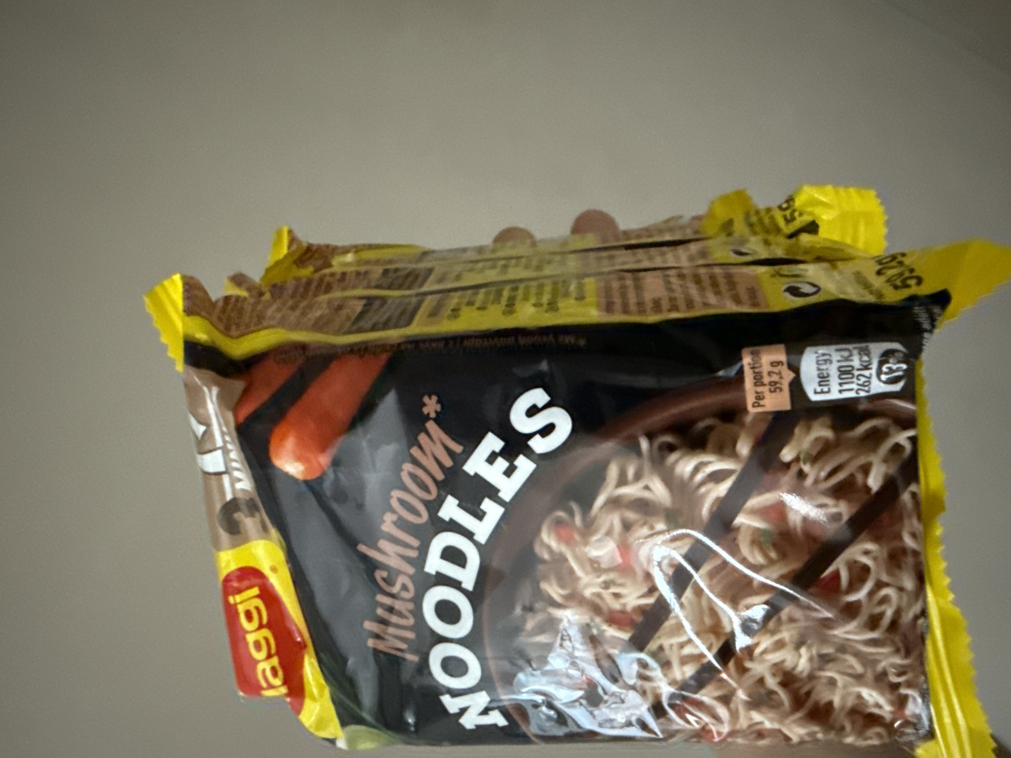 This is a close-up, slightly angled shot of a package of Maggi brand mushroom-flavored instant noodles. The packaging is predominantly black with white text that reads "Mushroom NOODLES." There is a yellow border along the top and right edges of the package, with some text and nutritional information visible. A red and yellow "Maggi" logo is visible on the left side of the packaging. The noodles themselves are visible through a clear plastic window on the front of the package, appearing as tightly coiled strands. The lighting appears to be artificial and the background is a plain, light gray surface, suggesting an indoor setting. There are no people or any specific location cues beyond the product itself. The visible text includes "Maggi," "Mushroom* NOODLES," "Με γευσή μανιταρι" (which translates to "with mushroom flavor" in Greek), "Per portion 59,2 g," and "Energy 1100 kJ 262 kcal." There are also some numbers like "13" and "59.2g" visible. The overall impression is a product shot, likely for identification or informational purposes.