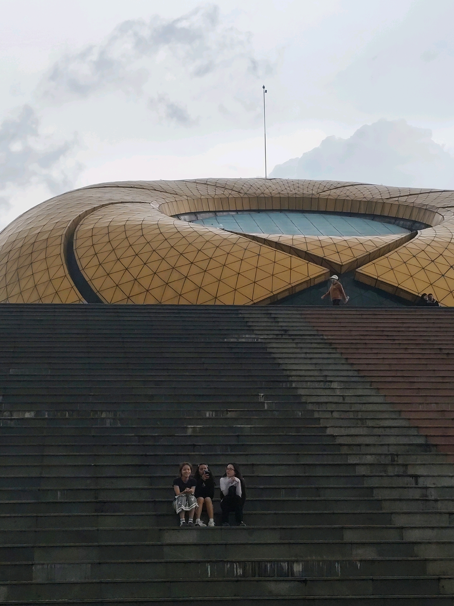 The image shows the entrance to a large, golden, dome-shaped building, which is likely the Da Lat Exhibition Center, or a similar modern structure, in Da Lat, Vietnam. The building's roof is made of a geometric, tessellated pattern. In front of the building is a very wide and long set of dark grey stone stairs.
On the upper part of the stairs, a person in an orange shirt and a white hat is walking. Further back, another person is visible. On the lower section of the stairs, three young women are sitting. One woman on the left is wearing a black shirt and a patterned skirt. The woman in the middle is wearing a black shirt and black pants. The woman on the right is wearing a white shirt and black pants. They appear to be in a casual setting, possibly enjoying the view or waiting for someone.
The sky is overcast with soft, white clouds. The overall impression is one of a large public space, likely a tourist attraction or a venue for events, with people enjoying a leisurely moment.