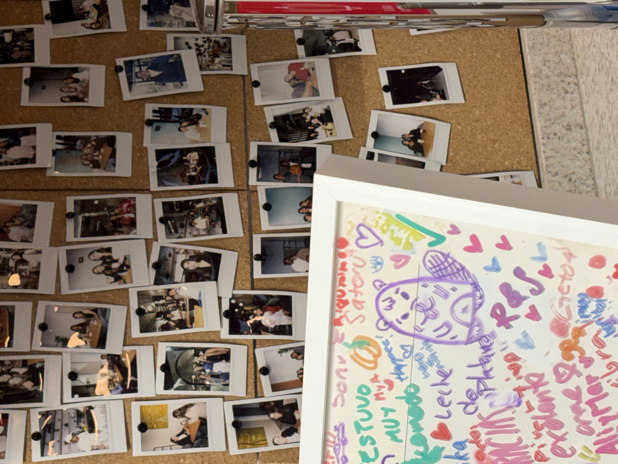 A corkboard covered with instant photos is partially visible in the upper left of the image. The photos are pinned to the board with black pushpins and are arranged in rows and columns. A white framed picture with colorful handwritten messages and doodles obscures part of the corkboard in the lower right. The messages are in different colors and appear to be written by multiple people. There is an indiscernible object on top of the corkboard. The background is a pale, neutral color and is out of focus. The location is Puebla, Mexico.