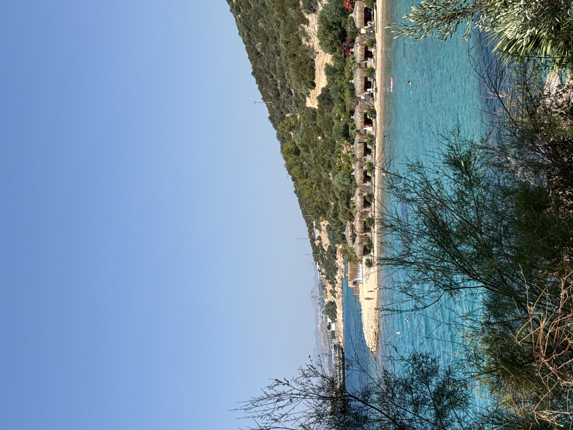 The image depicts a serene beach scene in Bodrum, Turkey. The foreground is dominated by the lush green foliage of trees and shrubs, partially obscuring the view. In the midground, a picturesque bay unfolds with crystal-clear turquoise waters. Along the shoreline, a stretch of golden sand is lined with numerous thatched-roof cabanas, creating a resort-like atmosphere. Several beach chairs are placed in front of these cabanas. Two individuals are visible on the sandy beach, and a few more are enjoying the water, with one person floating on a pink inflatable mattress. In the background, a moderately sloped hill is covered with dense greenery and scattered buildings, suggesting a town or village nestled on the hillside. A clear, cloudless blue sky stretches across the upper portion of the frame. The overall impression is one of a tranquil and inviting coastal destination.