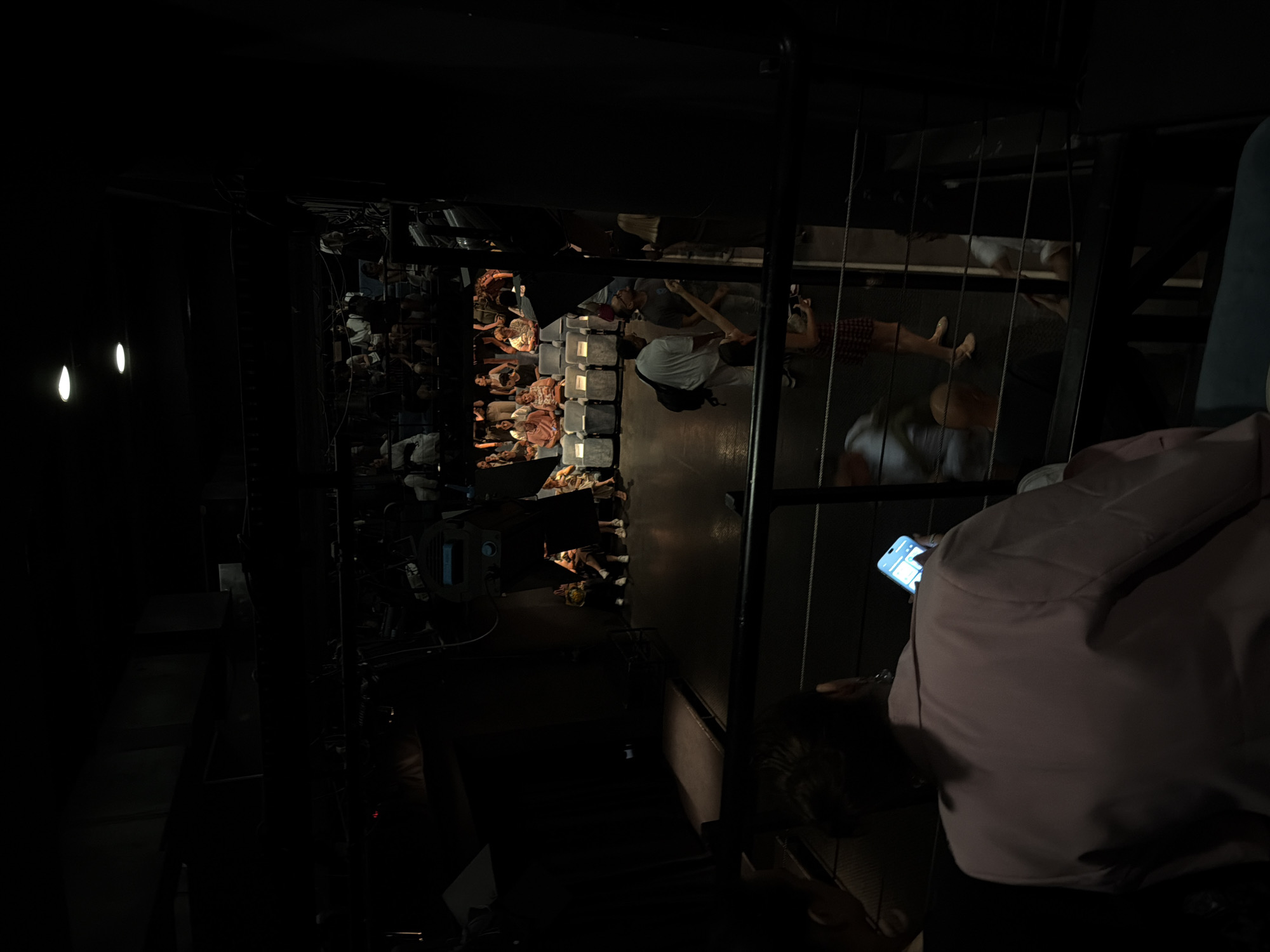 In Zvezdara Urban Municipality, Serbia, a dimly lit indoor space, likely a theater or performance venue, is captured. The perspective is from an elevated vantage point, looking down onto a seating area where an audience of approximately 30-40 individuals is gathered. The audience members are seated in rows of chairs, some looking towards an unseen stage. The lighting is subdued, with overhead spotlights casting pools of light onto the stage area, leaving much of the venue in shadow. In the foreground, a person's back is visible, facing away from the camera and towards the audience, holding a smartphone. Another person, also partially visible, is seated to the left. The overall atmosphere is one of anticipation or quiet observation before an event.
