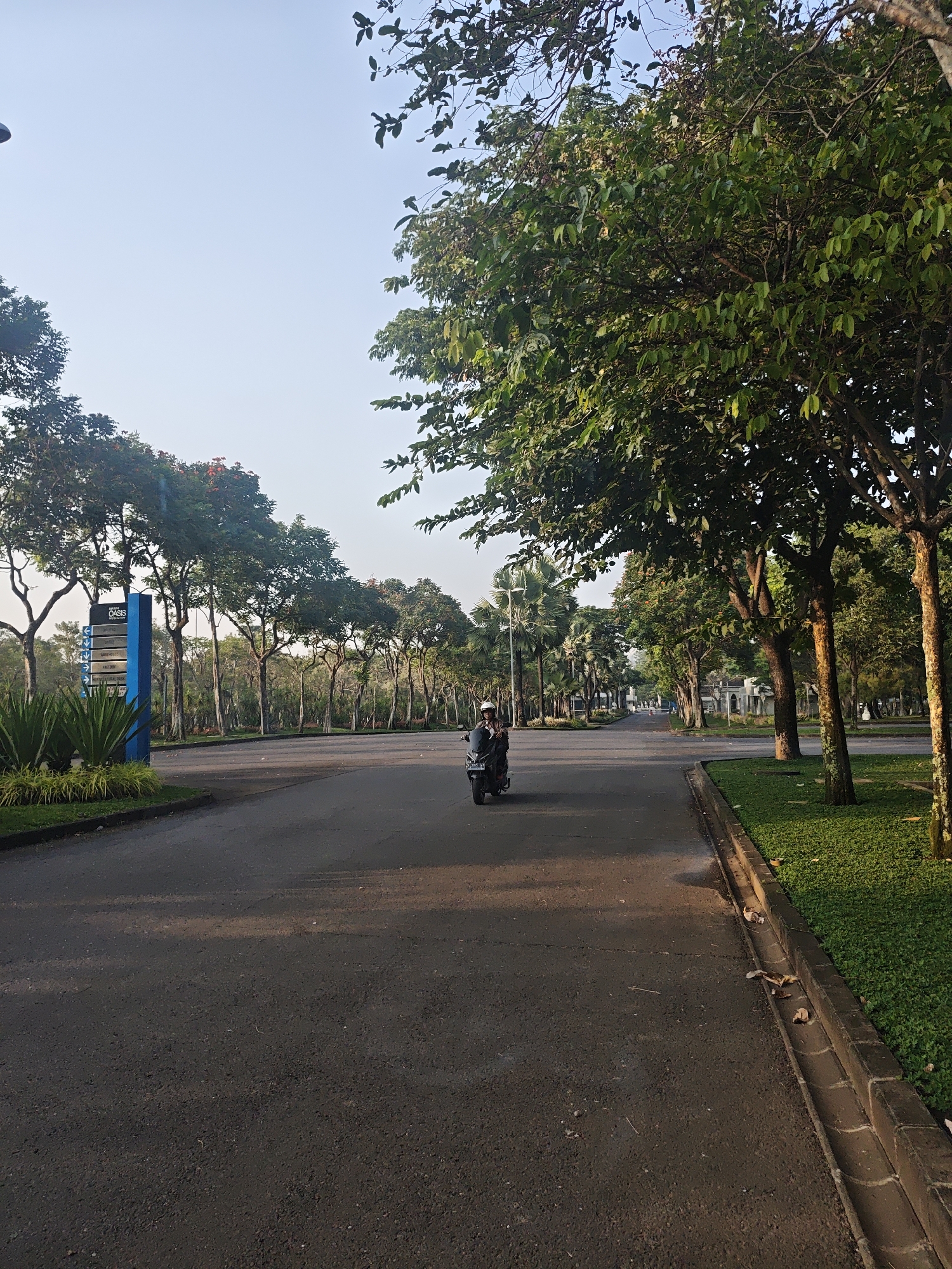 The image shows a paved road with a motorcycle rider in the distance, traveling away from the camera. The road is lined with trees and manicured grass on the right side. A blue signpost with white text and arrows is visible on the left side, indicating directions to various locations within "OASIS". The sky is clear and blue, suggesting it is daytime, likely early morning or late afternoon due to the soft lighting. The environment appears to be a well-maintained, possibly residential or commercial area, with a focus on greenery. The image was captured in the city of Bae, Indonesia, given the provided context.