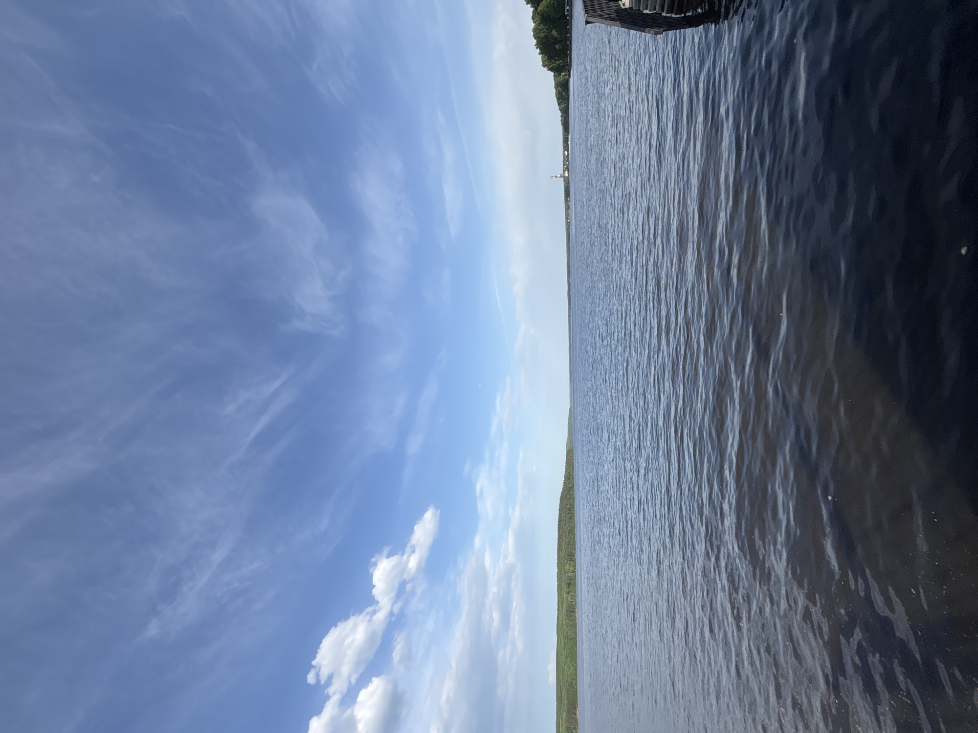 The image shows a body of water, possibly a lake or river, with a clear blue sky above. The water is dark and slightly rippled. A line of trees and vegetation is visible along the shore in the middle of the image. In the distant background, a light-colored structure can be seen. Part of a dock or other wooden structure is visible at the top right of the image. The location is Unknown, Unknown.