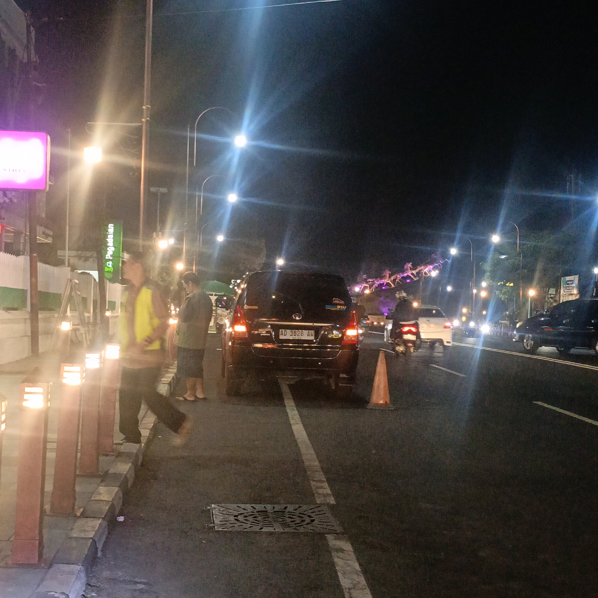 The image depicts a street scene at night in Boyolali, Indonesia, characterized by artificial lighting from streetlights, illuminated signs, and decorative fixtures. A dark-colored SUV with license plate "AD 1828 AW" is parked on the right side of the road, facing away from the viewer. A traffic cone is positioned behind the SUV. To the left of the SUV, a person wearing a bright yellow vest and dark pants is standing on the sidewalk. Further to the left, another person is visible, facing away from the camera. Decorative street lamps are lining the sidewalk, emitting a warm glow. In the background, there are more streetlights, trees, and what appear to be decorative lights forming a structure across the road. The overall ambiance is that of a well-lit urban street at night.