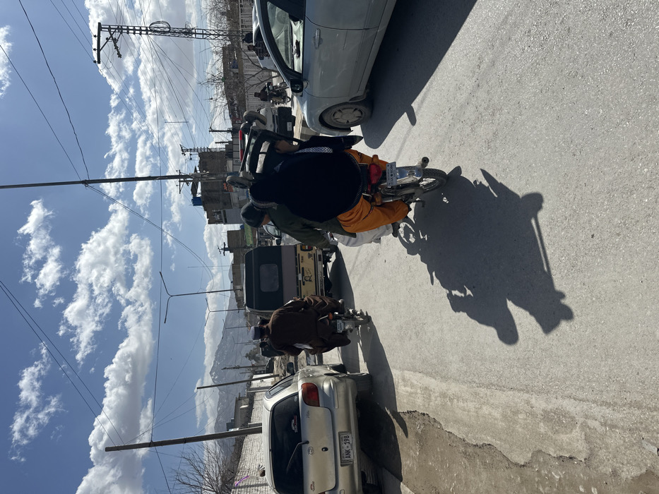 The image depicts an outdoor street scene in Quetta City Tehsil, Pakistan, under a bright blue sky with scattered white cumulus clouds. Strong sunlight casts long, distinct shadows of people and vehicles onto the asphalt road.
In the immediate foreground, two motorcycles are traveling away from the viewer. The motorcycle on the right carries two individuals: a driver wearing a green top and white lower garment, and a passenger enveloped in a dark blue/black garment over orange lower clothing. This passenger is transporting a white and red object, consistent with a child seat or a small portable chair, on their back. The motorcycle on the left is operated by a single individual dressed in brown traditional attire.
Further down the road, a beige three-wheeled auto-rickshaw is visible. On the left side of the frame, a silver Toyota Vitz hatchback (license plate ANK-198) is parked on the shoulder. On the right, a silver sedan is present. The asphalt road surface exhibits wear, and a rough dirt shoulder is visible on the left.
The background features low-rise buildings on both sides of the street. Numerous utility poles with complex networks of overhead power lines and streetlights are visible. A prominent tall metal lattice tower stands on the far right. In the distant background, light-colored mountains are visible under the clear sky. A low wall topped with barbed wire or razor wire runs along the left side of the road, adjacent to the parked vehicle.