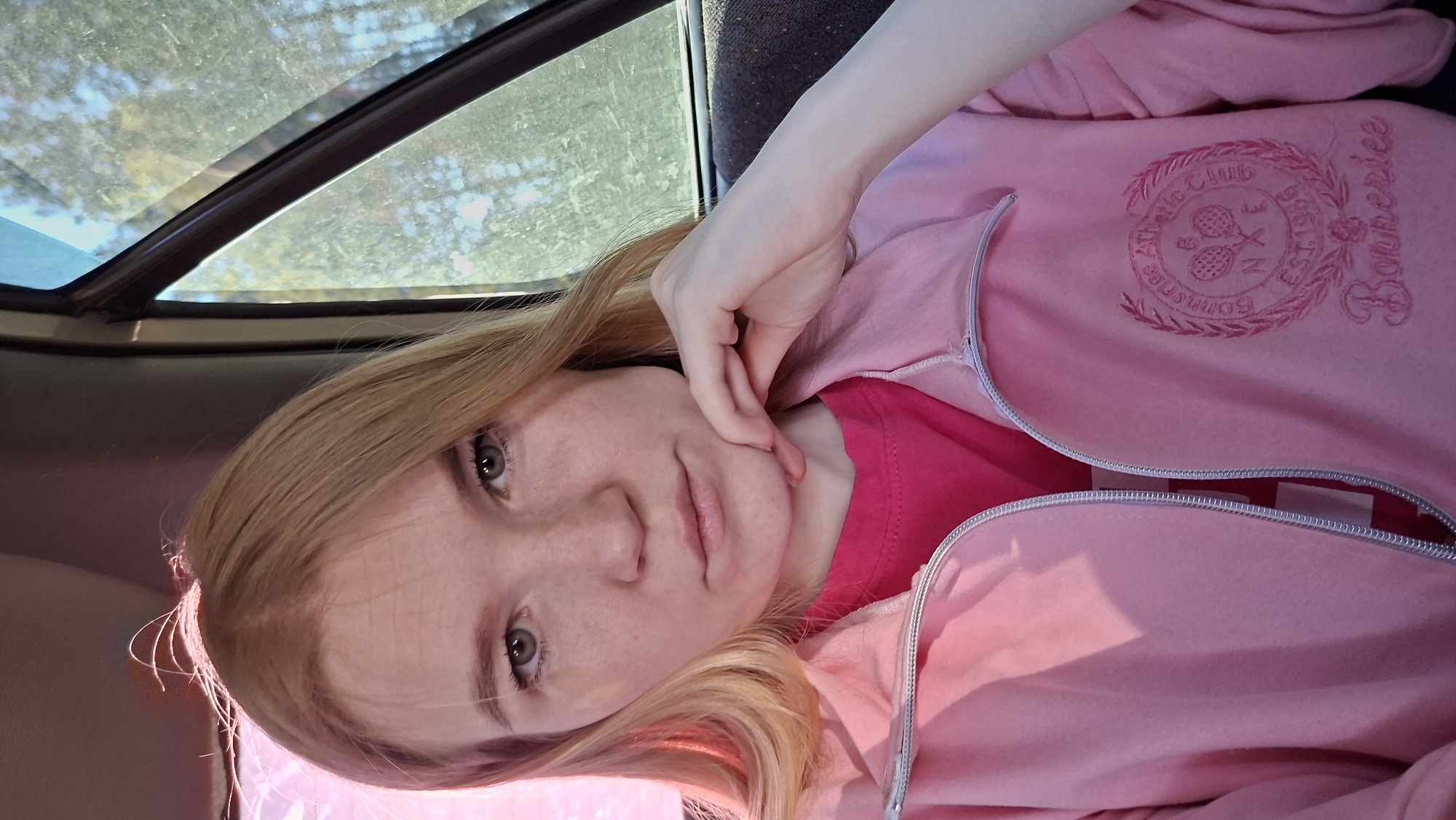 A fair-skinned person with light brown hair is seated inside a vehicle, wearing a pink zip-up sweatshirt with a red shirt underneath. The sweatshirt has an embroidered design reading "Bonnerice Athletic Club Est 1998." The person's hand is touching their chin. A sunroof is visible above. The specific location within the city of Chaikovsky, Russia, cannot be determined from the image.