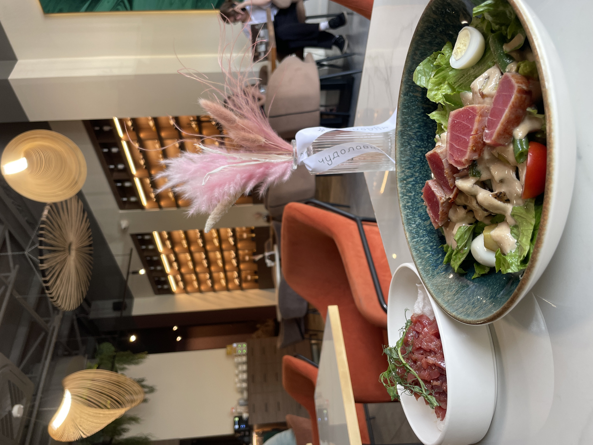 The image captures a dining scene in a modern restaurant, presented with a 90-degree counter-clockwise rotation. On a light-colored table in the foreground, two artfully presented dishes are visible. One is a salad in a dark-rimmed bowl, featuring seared tuna slices, fresh green lettuce, halved quail eggs, tomato wedges, and a creamy dressing. Beside it, a smaller white bowl contains a dish that appears to be tuna tartare, garnished with green microgreens. Between the dishes sits a glass vase containing an arrangement of pink and beige feathery ornamental grasses. A white ribbon wrapped around the vase prominently displays the text "ЧУДОЛОВ" (CHUDOLOV).
The background reveals the restaurant's contemporary interior design. There are vibrant orange upholstered chairs, contributing a pop of color to the otherwise neutral palette. Dark wooden paneling or shelving units, illuminated from within, create a grid pattern on the walls. Unique, sculptural light fixtures, designed with a woven or spiral pattern, hang from the ceiling. A large reflective surface, likely a mirror or polished wall/ceiling, shows the inverted reflection of a person seated at a table further back, dressed in dark clothing and a white top. The general brightness suggests it is daytime. This setting is indicative of an upscale casual dining experience in Kazan, Russia.