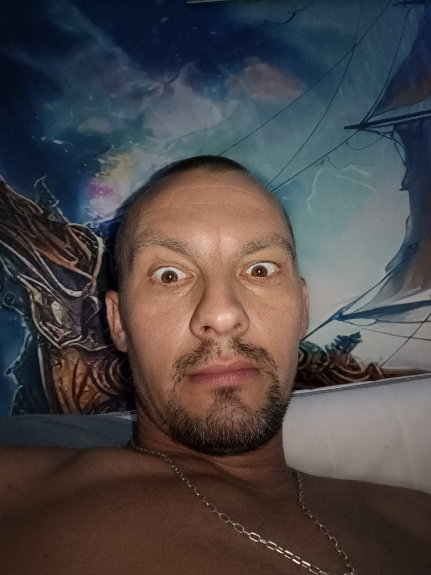 A man with a shaved head and a goatee is looking directly at the camera with wide eyes. He is wearing a silver chain around his neck. The background appears to be a mural or tapestry featuring a dark, stormy sky with lightning and the silhouette of a sailing ship. To the left of the man, a portion of a dark, armored figure or creature is visible. The lighting is dim, creating a dramatic effect. This image was taken indoors, likely in a residential setting, and there is no specific indication of its location in Bolesławiec, Poland.