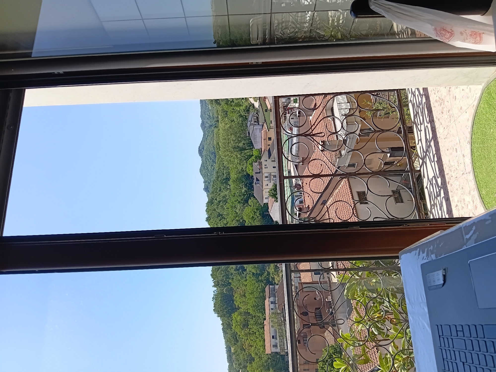 The image captures a view from a balcony in Vallesaccarda, Italy. The foreground shows a portion of a laptop covered in plastic wrap, placed on a white surface. Beyond the laptop, a decorative black metal railing lines the balcony. Through the railing, an outdoor scene unfolds with red-tiled roofs of houses, some with balconies and windows. Lush green trees and vegetation cover the landscape, leading up to rolling hills in the background under a clear blue sky. The reflection on a glass surface on the right side of the image shows a chair and a table on what appears to be another balcony or outdoor seating area. The overall impression is one of a quiet, residential area in a hilly landscape.