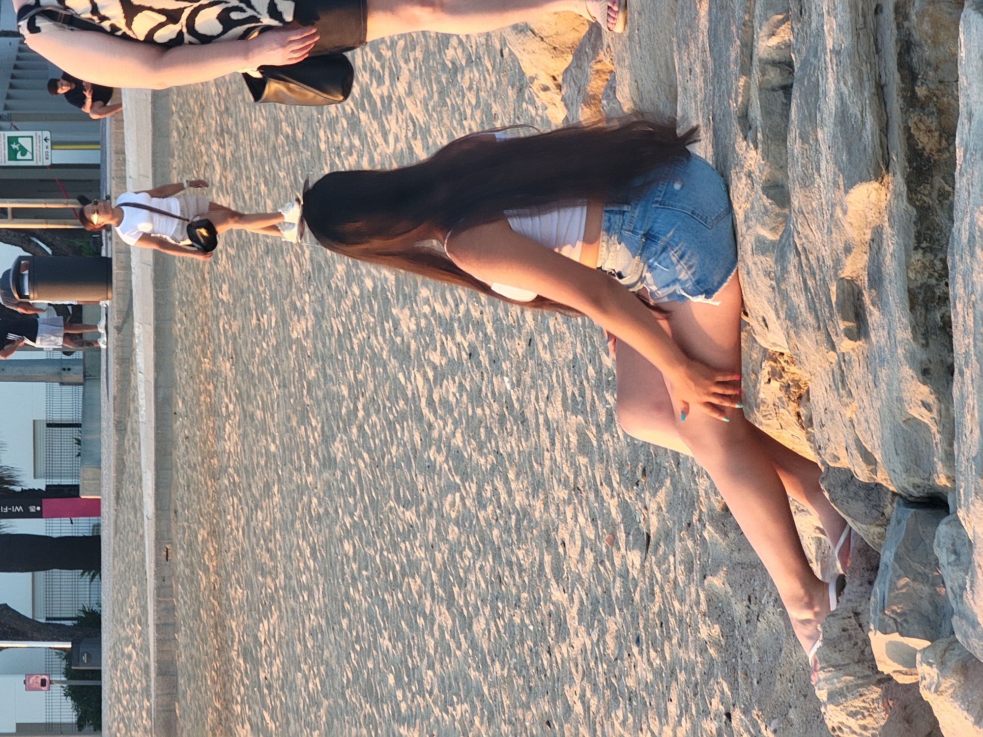 The image depicts a sunny day on a sandy beach in Quarteira, Portugal. In the foreground, a young woman with long, dark hair sits on large rocks, facing away from the camera. She is wearing a white crop top and denim shorts. Her legs are extended, and she is wearing white flip-flops. Her right hand, resting on a rock, shows colorful fingernails. To her right, partially visible, is another person's legs and arm, wearing a black and white patterned garment and black shorts, along with a black handbag.
In the middle ground, a woman walks across the sand. She has short, dark hair, sunglasses, and is wearing a white t-shirt, beige shorts, and white sneakers. She carries a black purse over her shoulder.
In the background, other people are visible further up the beach, near a walkway with metal fencing. One person is wearing a black t-shirt and grey shorts, and another is seated. The environment includes sand, rocks, and a paved area with a metal fence and some vegetation. The overall lighting suggests it is late afternoon or early evening.