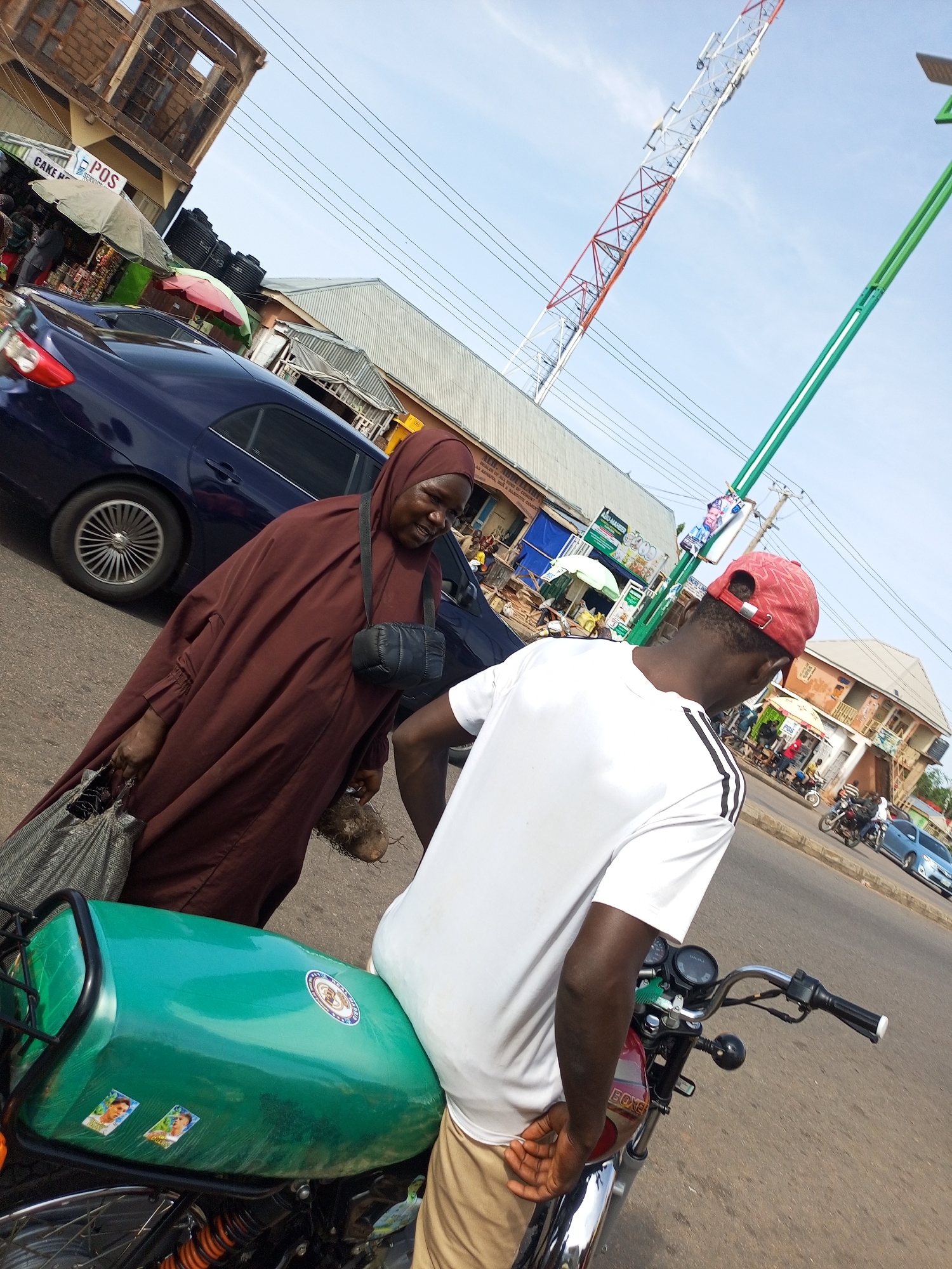 The image depicts a street scene in Tempure, Nigeria, on a bright, sunny day. In the foreground, a man wearing a white t-shirt and a red baseball cap is seated on a green motorcycle. His back is to the camera, and his right arm is resting on the motorcycle's fuel tank. To his left, a woman in a dark brown hijab and dress is walking, carrying a large bag in her left hand and what appears to be a root vegetable in her right. Her gaze is directed towards the man.
Behind them, a dark blue car is parked on the side of the road. The background reveals a bustling street with various shops and buildings. Umbrellas are set up in front of some of the stalls, suggesting a marketplace. A prominent red and white telecommunications tower rises into the sky in the upper right portion of the image, indicating the urban setting. Several other people and motorcycles are visible in the distance, adding to the lively atmosphere of the street. The road surface is asphalt, and power lines crisscross overhead.