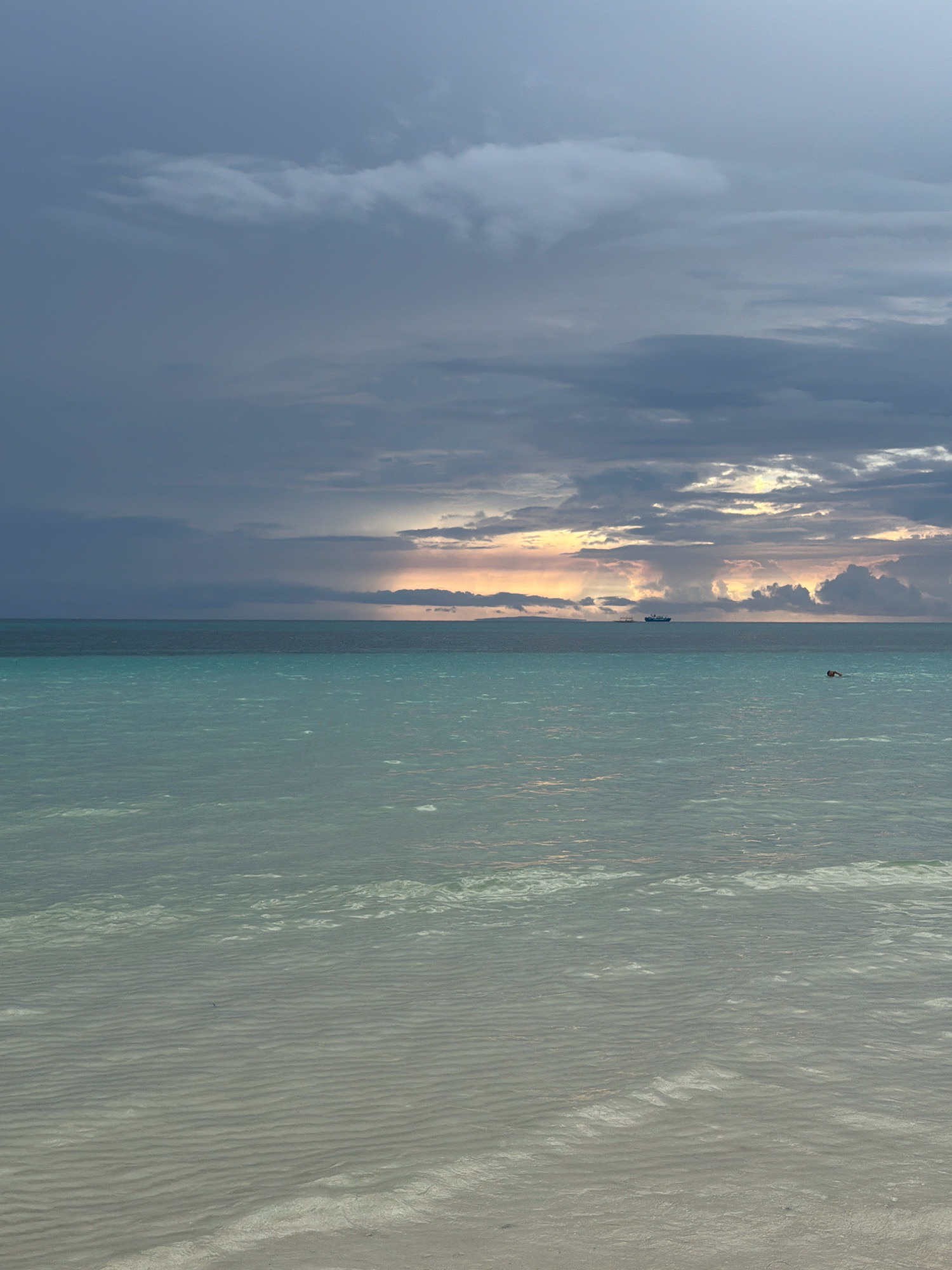 The image depicts a serene seascape under a dramatic, cloudy sky. The horizon is sharply defined, separating the turbulent, multi-hued sky from the tranquil ocean. The sky is filled with dark, brooding clouds, interspersed with patches of lighter grey and hints of golden light peeking through near the horizon, suggesting either sunrise or sunset.
The ocean itself is divided into two distinct bands of color. The upper band, closer to the horizon, is a deep turquoise, gradually transitioning into a lighter, almost translucent aquamarine closer to the foreground. The water's surface is rippled, with faint patterns formed by the receding tide on the sandy seabed visible in the shallows.
In the distance, a large ship or ferry is visible on the horizon, a solitary object against the vastness of the sea and sky. To the right of the ship, a single person is swimming or floating in the water. The foreground reveals a sandy beach, mostly submerged by the shallow water, showing the gentle undulations of the sand beneath the surface. The overall impression is one of peaceful isolation and the raw beauty of nature.
Based on the visual characteristics of the ocean, sky, and the presence of a beach with shallow, clear water, this scene is consistent with coastal areas in Malay, Philippines, particularly locations like Boracay Island, known for its stunning beaches and clear waters.