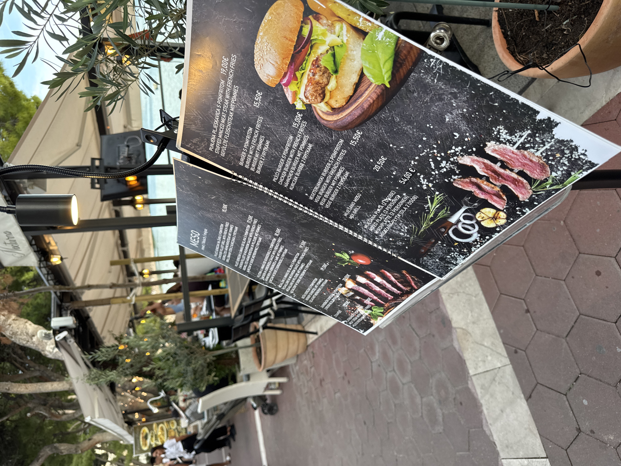 An outdoor dining menu stands at an angle on a tiled patio, suggesting a restaurant setting in Makarska, Croatia, likely near the coast given the clear blue sky and distant water visible behind the trees and awnings. The menu is a dark-colored, glossy tri-fold with images of food, including a burger and grilled meat. Text on the menu is in multiple languages, with prices indicated in Euros. Several tables and chairs for diners are visible in the background, along with potted plants and trees providing shade. People are seen in the background, including a woman walking away from the camera in a white top and black pants. The scene is well-lit, likely during daytime, with some artificial lights visible on the restaurant's awning.