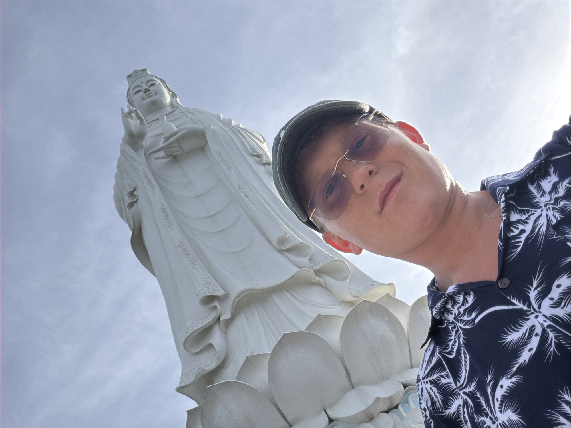 A low-angle, medium shot captures a person in front of the Lady Buddha statue at the Linh Ung Pagoda, in Đà Nẵng, Vietnam. The person, positioned on the right side of the frame, wears a patterned dark blue short-sleeve button-up shirt with white palm trees, tinted sunglasses, and a dark green hat. Their face is turned towards the camera. The Lady Buddha statue stands tall in the background, on the left side of the image. The statue is white, depicting a female figure in flowing robes. The sky behind them is a light, overcast blue. The statue is standing on large lotus petals.