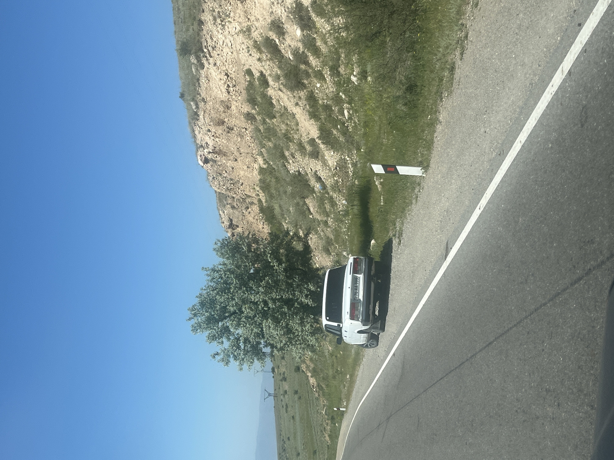 The image, oriented correctly, depicts a roadside scene on a bright, sunny day. The primary subject is a white sedan, possibly a Lada, parked on the right shoulder of a paved road. The car is seen from its rear-left side, facing away from the camera. Its license plate, clearly visible, reads "E 76 AA 999".
To the left of the road, a steep, rocky hillside rises, covered sparsely with green patches of grass and shrubs. A large, leafy green tree stands between the parked car and the base of the hillside, casting a shadow onto the ground. Further in the distance, to the left and beyond the hillside, a vast, undulating landscape with more greenery and distant hills or mountains under a clear blue sky is visible.
The road itself is asphalt, featuring a distinct solid white line marking its right edge. A small white and red/black striped roadside marker post is situated near the car, further down the road. The sky is a vibrant, clear blue, indicating fair weather. The strong shadows cast by the car and the hillside suggest that the sun is high, likely mid-morning to afternoon. No people are visible in the scene. The overall setting is rural or mountainous, hinting at a journey or a stop along a scenic route.