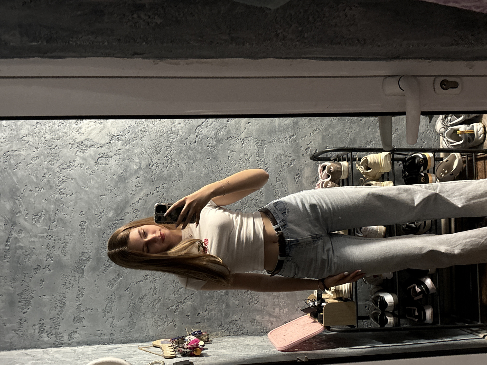 A young woman with long blonde hair is taking a selfie in a mirror. She is wearing a white t-shirt and blue jeans with a black belt. The background shows a textured gray wall, a shoe rack with various shoes, and a windowsill. The location is implied to be Panagyurishte, Bulgaria.