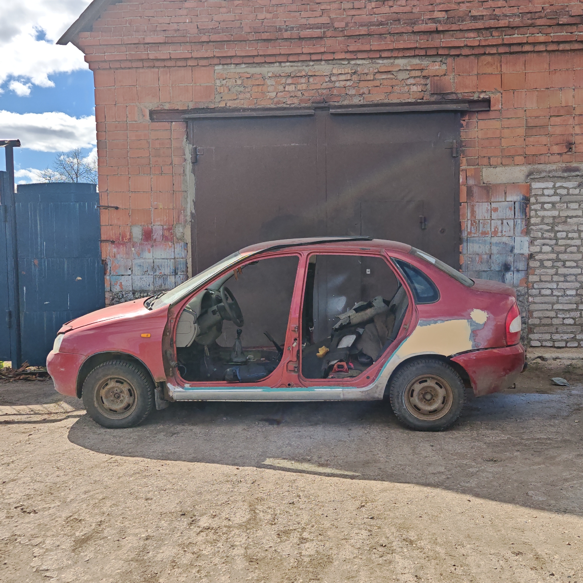 A red Lada car, stripped of its doors, stands in front of a brick garage and a blue metal gate. The garage has two large, dark-colored metal doors, and some of the brickwork on the garage has been patched. The car's interior is visible, revealing the steering wheel, dashboard, and seats, some of which are covered with what appears to be gray material. The exterior of the car shows signs of wear, with patches of body filler on the rear quarter panel. The ground is dirt and gravel. The sky is partly cloudy. The location is Rostov, Russia.