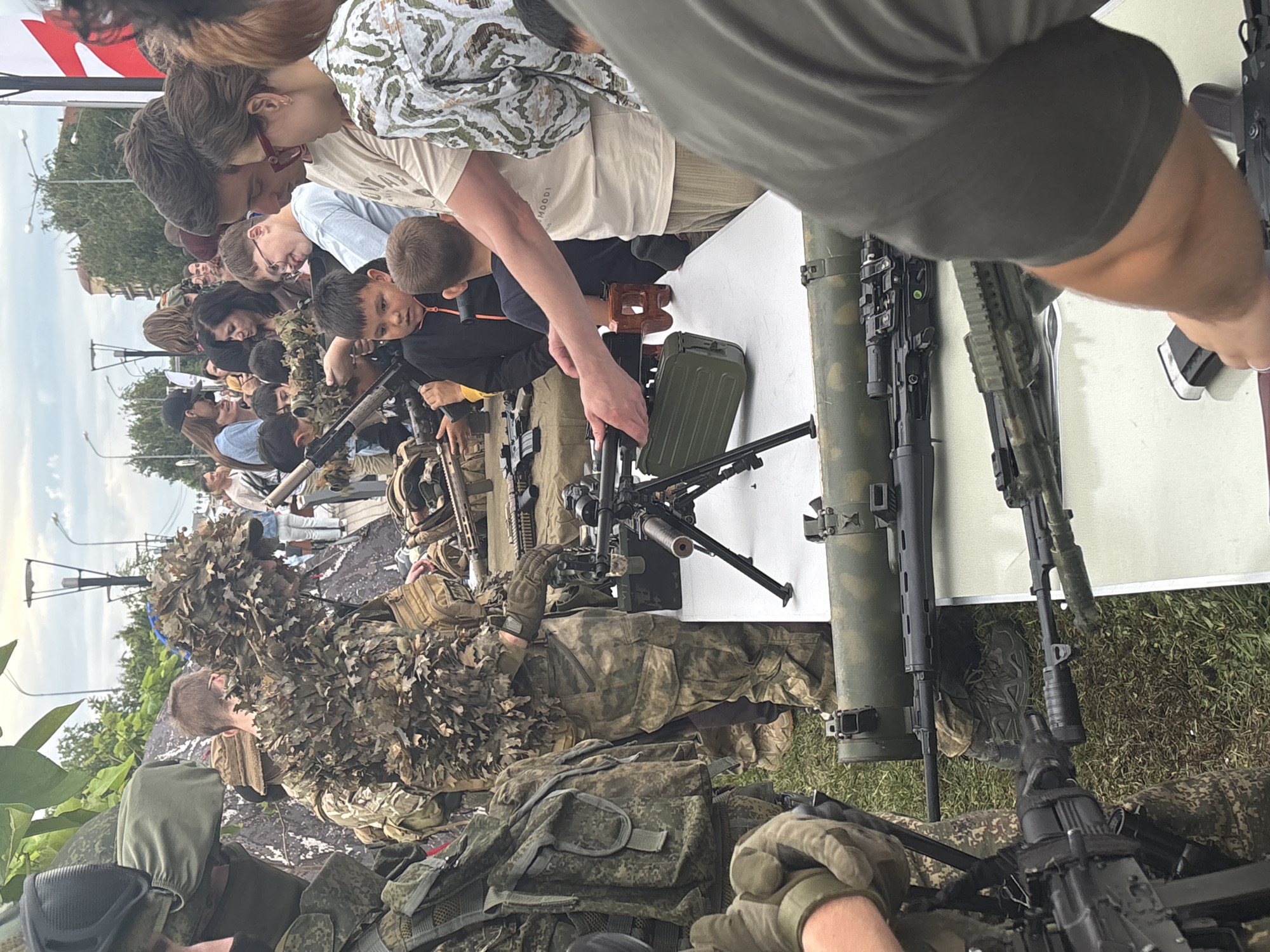 The image captures a gathering of people, including adults and children, in an outdoor setting. Several individuals are dressed in camouflage clothing, with one person wearing a ghillie suit. Various firearms and military equipment are on display, including what appears to be a machine gun mounted on a tripod, a rocket-propelled grenade launcher, and several rifles. The scene suggests an event related to military hardware or perhaps a survivalist or reenactment gathering.
The people in the image show a range of engagement, with some observing the equipment and others interacting with it. The atmosphere appears to be one of curiosity and interest, particularly among the younger attendees. The presence of both adults and children indicates a family-friendly event, or at least one where children are present.
The setting is outdoors, with grassy terrain and some vegetation visible. In the background, trees and structures suggest an urban or semi-urban environment. The lighting indicates it is daytime, likely afternoon given the angle of the shadows. The weather appears to be overcast or hazy. There are no discernible text elements in the image. The location context of Astrakhan, Russia, might imply a local event or exhibition.