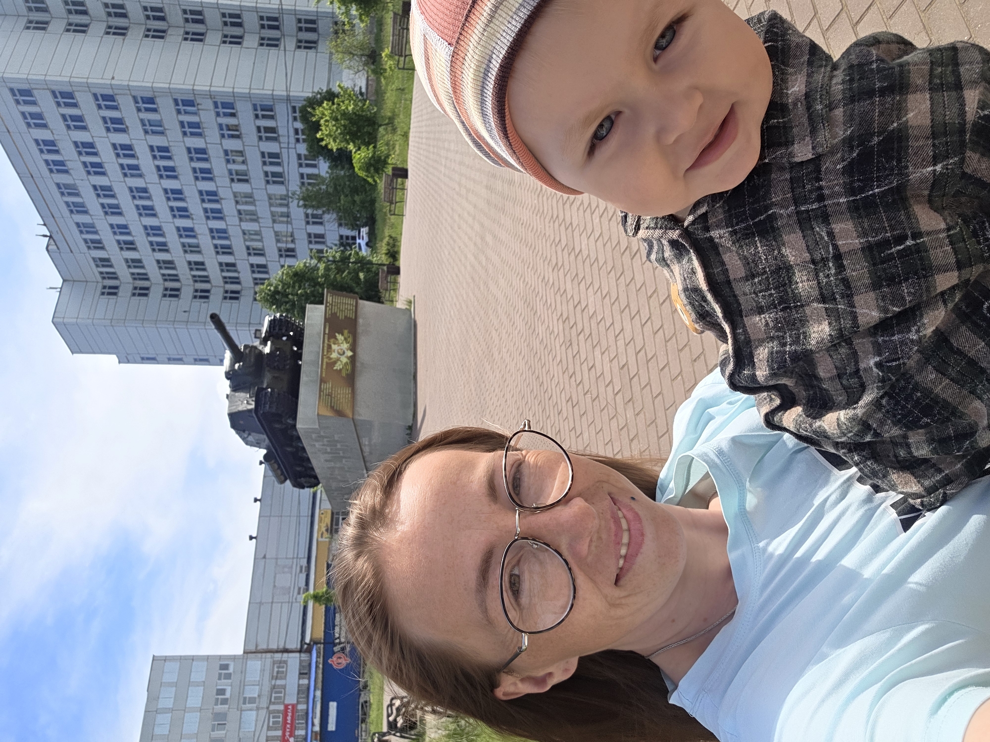 A selfie captures a woman and a young child in an outdoor setting in Chaikovsky, Russia, on a bright, sunny day. The woman, with long brown hair and round eyeglasses, is in the foreground on the right, smiling warmly at the camera. She is wearing a light blue t-shirt and a silver necklace, and has freckles visible on her face. To her left, the child, likely a baby or toddler, is held up and looking towards the camera with a gentle smile. The child wears a striped beanie hat with earthy tones and a plaid shirt in dark green and brown hues.
In the background, a paved plaza with interlocking bricks extends behind them. To the left, a large, multi-story building with numerous windows dominates the upper background. Further back and slightly to the left, a dark-colored tank is mounted on a pedestal, appearing to be a memorial. A plaque with Cyrillic text and an emblem is visible on the monument base next to the tank, though the text is not clearly legible. Scattered trees and green foliage are visible between the buildings and the monument. Another building with a blue facade and a visible sign reading "WWW.KAB..." and "ЗАБОТА" (ZABOTA) in Cyrillic can be seen in the distance, partially obscured. The sky is clear blue with sparse white clouds, indicating pleasant weather. Both individuals appear happy and content, enjoying their time outdoors.