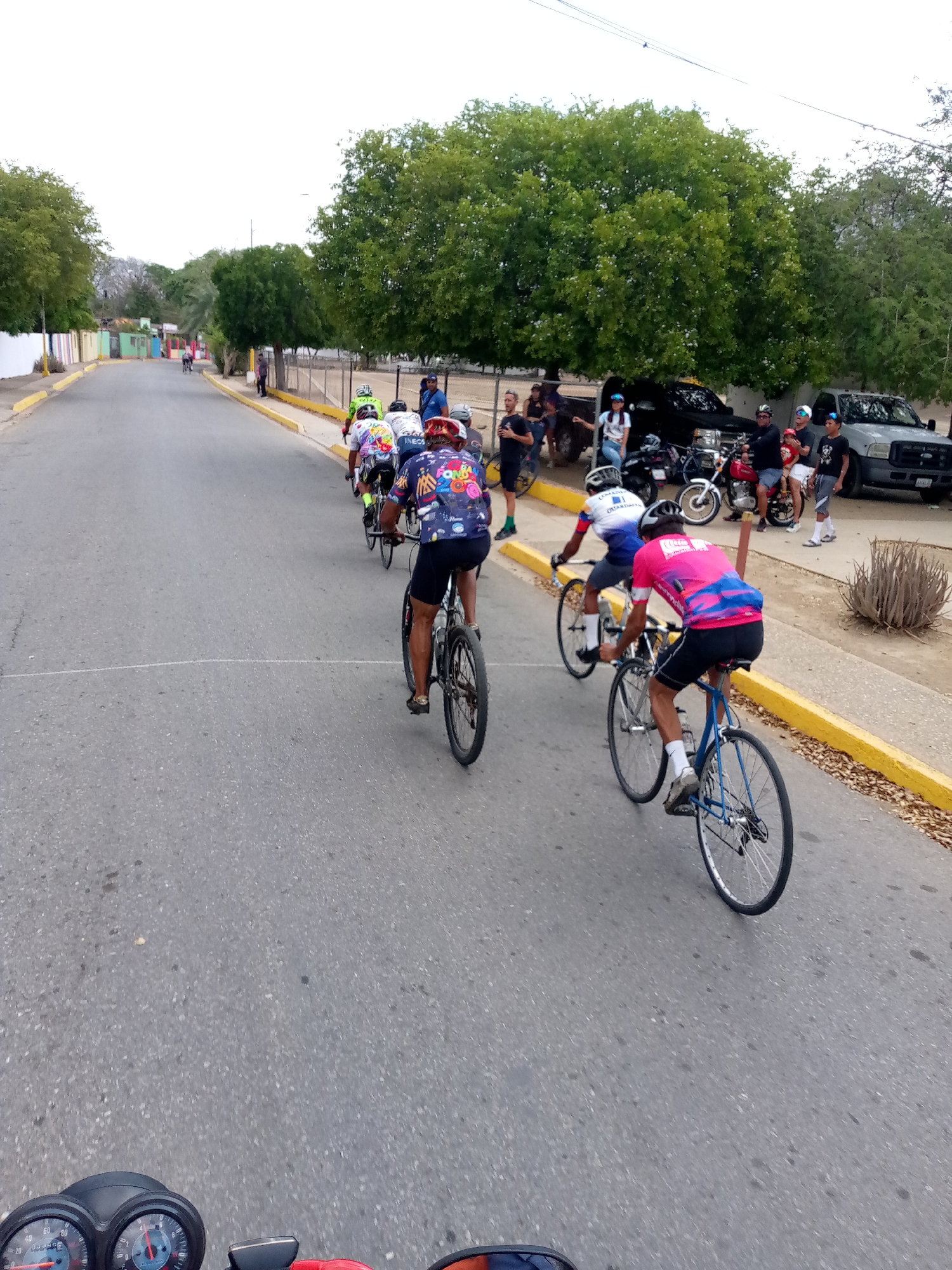 The image captures a bicycle race on a paved road in what appears to be Guatacaral, Venezuela. A group of cyclists, wearing colorful jerseys and helmets, are riding in formation, with some spectators standing on the side of the road. Parked motorcycles and cars line the side of the road where the spectators gather. The surroundings include green trees, buildings, and a yellow curb. The viewpoint is from a motorcycle following the race.