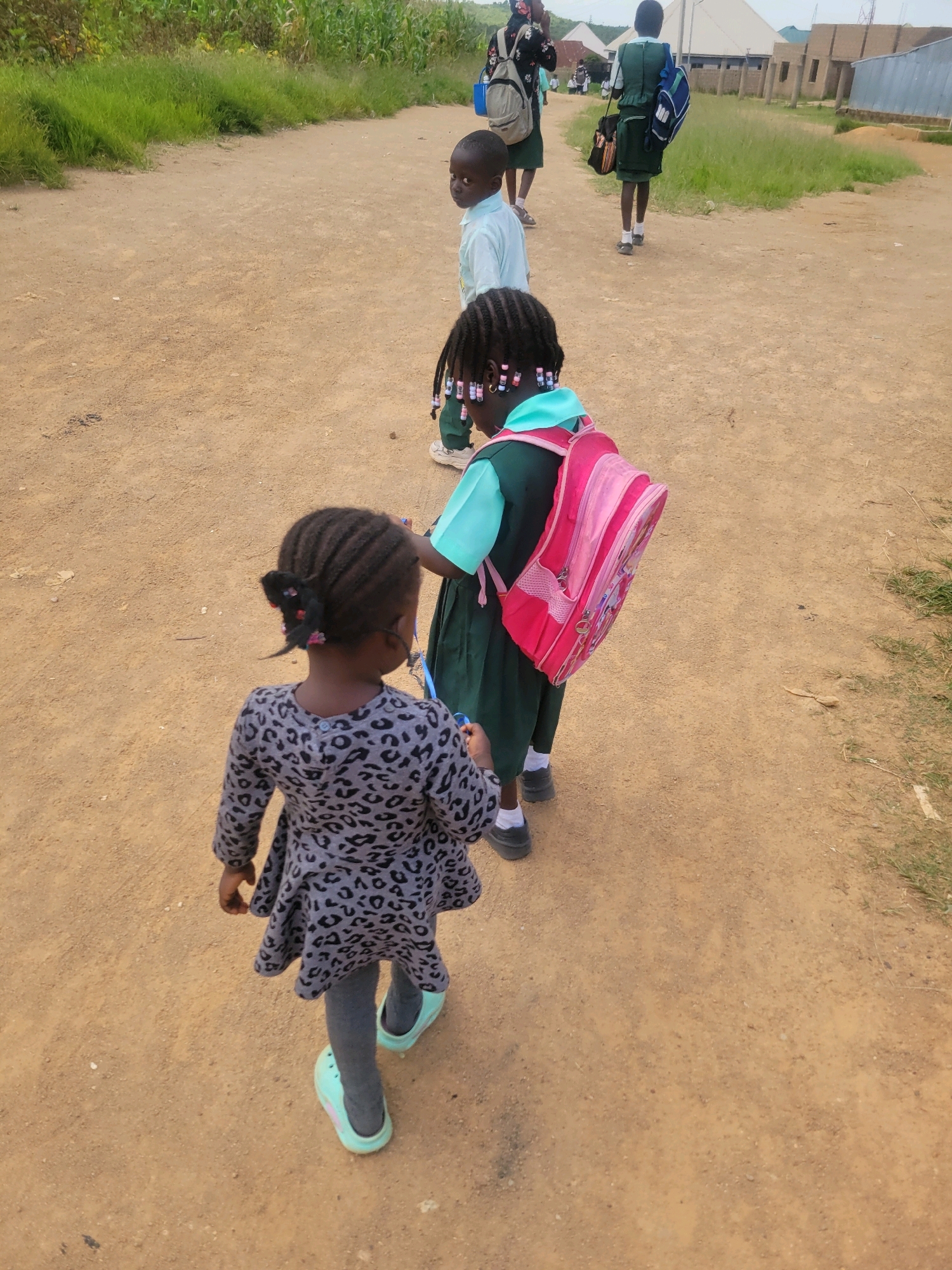In an outdoor, dusty environment, possibly in Jos, Nigeria, four young students are seen walking on a dirt path. The students are dressed in school uniforms, including green skirts and light blue shirts, and are carrying backpacks. The student in the foreground, a young girl with braided hair and a gray leopard-print dress over gray leggings and light blue shoes, is walking away from the camera. To her right, another student, a girl wearing a green skirt and a light blue top, carries a pink backpack adorned with a cartoon character. Further back, a boy in a light blue shirt is visible, and another student in a green uniform and a blue backpack is walking away from the camera. The background shows dry vegetation, including corn stalks, and some buildings in the distance. The lighting suggests it is daytime.