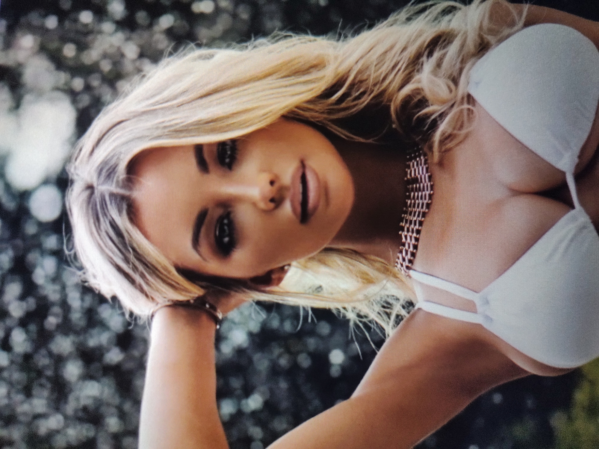 A blond woman is shown from the shoulders up, wearing a white bikini top and a gold chain necklace. She is looking at the camera with her head tilted slightly. Her left arm is bent at the elbow, with her hand resting on her head. The background is out of focus, showing trees and shrubs. The location is unknown.
