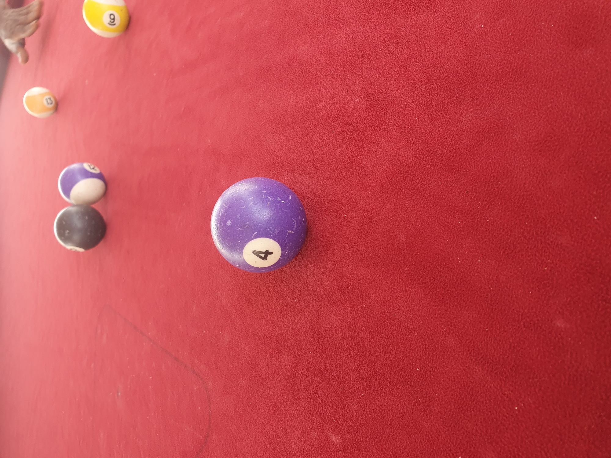 The image depicts a close-up view of a red pool table, likely in Lokoja, Nigeria, with several billiard balls scattered across the surface. In the foreground, a prominent purple ball with the number '4' clearly visible is centrally positioned. To the left and slightly behind it, a dark solid ball and a purple and white striped ball are visible. Further into the background, towards the upper right quadrant, a yellow and white striped ball with the number '13' and another yellow and white striped ball with the number '9' are present. A partial view of a person's hand, possibly in the act of playing or adjusting a ball, is visible in the top right corner of the frame. The red felt of the pool table exhibits a textured surface with a subtle sheen, and some dust particles are visible. The lighting suggests an indoor environment.