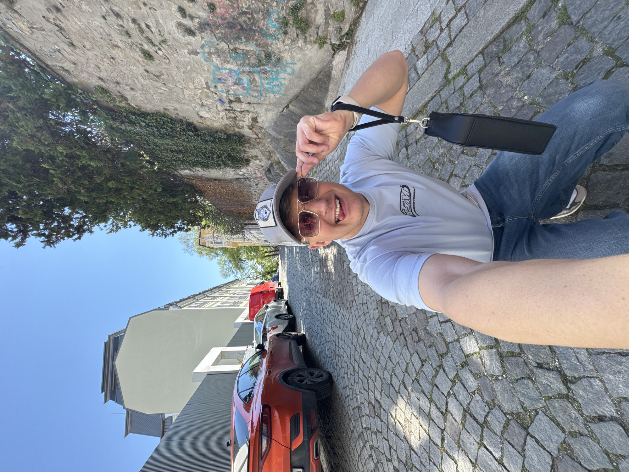 A man is taking a selfie on a cobblestone street lined with parked cars and buildings. The man, wearing a light-colored t-shirt, blue jeans, sneakers, sunglasses, and a baseball cap with a wolf emblem, is positioned at an angle to the camera. He is partially crouched or sitting, with the cobblestone street visible beneath him. The man is looking directly at the camera with his tongue sticking out and pointing to the brim of his baseball cap with his left hand.
The background shows a row of cars parked along the street, including an orange SUV closest to the camera, followed by a silver and then a red car. A gray building is located on the left, with the sky showing above. A stone wall, covered with graffiti and some vegetation, runs along the left side of the frame. The street appears to be in Plovdiv, Bulgaria, judging by architectural styles and the presence of cobblestone pavement.
The man is holding a small black bag or purse, with a strap attached, in his left hand. The image is taken during daylight hours, with bright sunlight illuminating the scene.