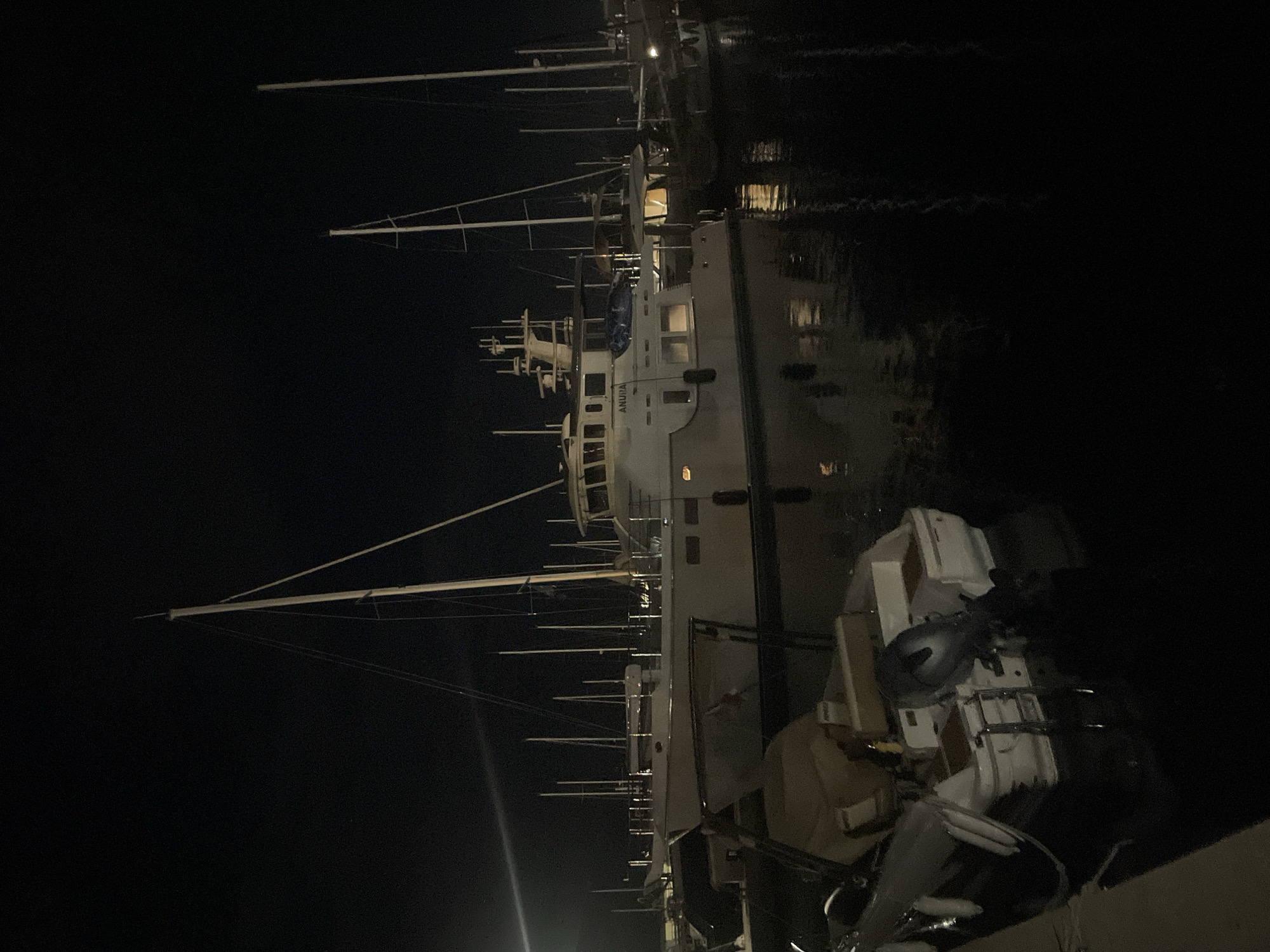 At night, a large white yacht named "ANURA" is docked in the city of Bar, Montenegro. The yacht is illuminated from within, casting a warm glow onto the dark water. Several other sailboats and smaller vessels are also docked nearby, their masts and rigging silhouetted against the night sky. The water reflects the lights of the boats and the city, creating a serene and tranquil atmosphere. The overall scene depicts a peaceful night at the harbor.