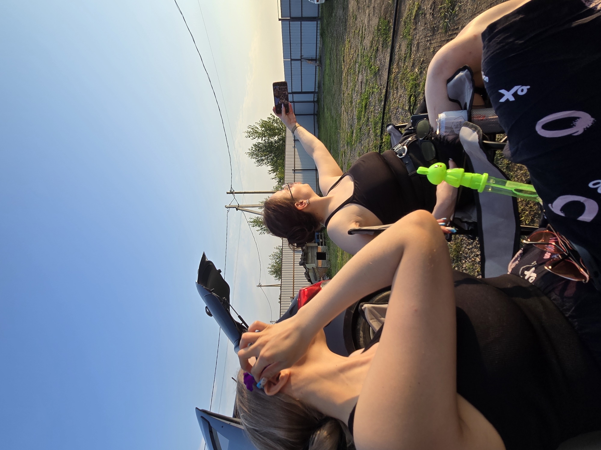 The image depicts two individuals seated in camping chairs outdoors. The woman on the left is facing away from the camera, with her hands near her head. The woman on the right is facing left and holding a smartphone, appearing to take a selfie. She is wearing glasses and a black tank top. A green bubble wand and a can of beverage are visible near her. The background shows a fence, trees, and a clear sky. The scene appears to be set in the vicinity of Ермолаево, Russia.