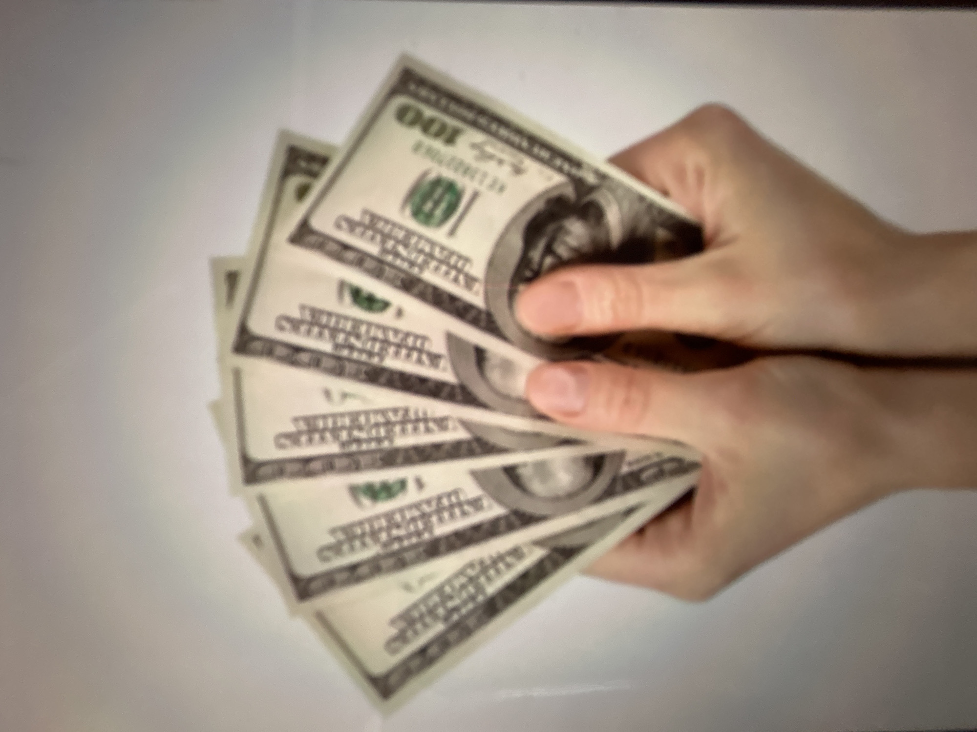 The image shows a pair of hands holding a fan of US one hundred dollar bills. The hands appear to be of a person with fair skin. The background is a plain, light-colored surface, possibly a wall or backdrop. The lighting is neutral, casting no strong shadows. There is no indication of the city of Gelendzhik, Russia, or any other specific location.