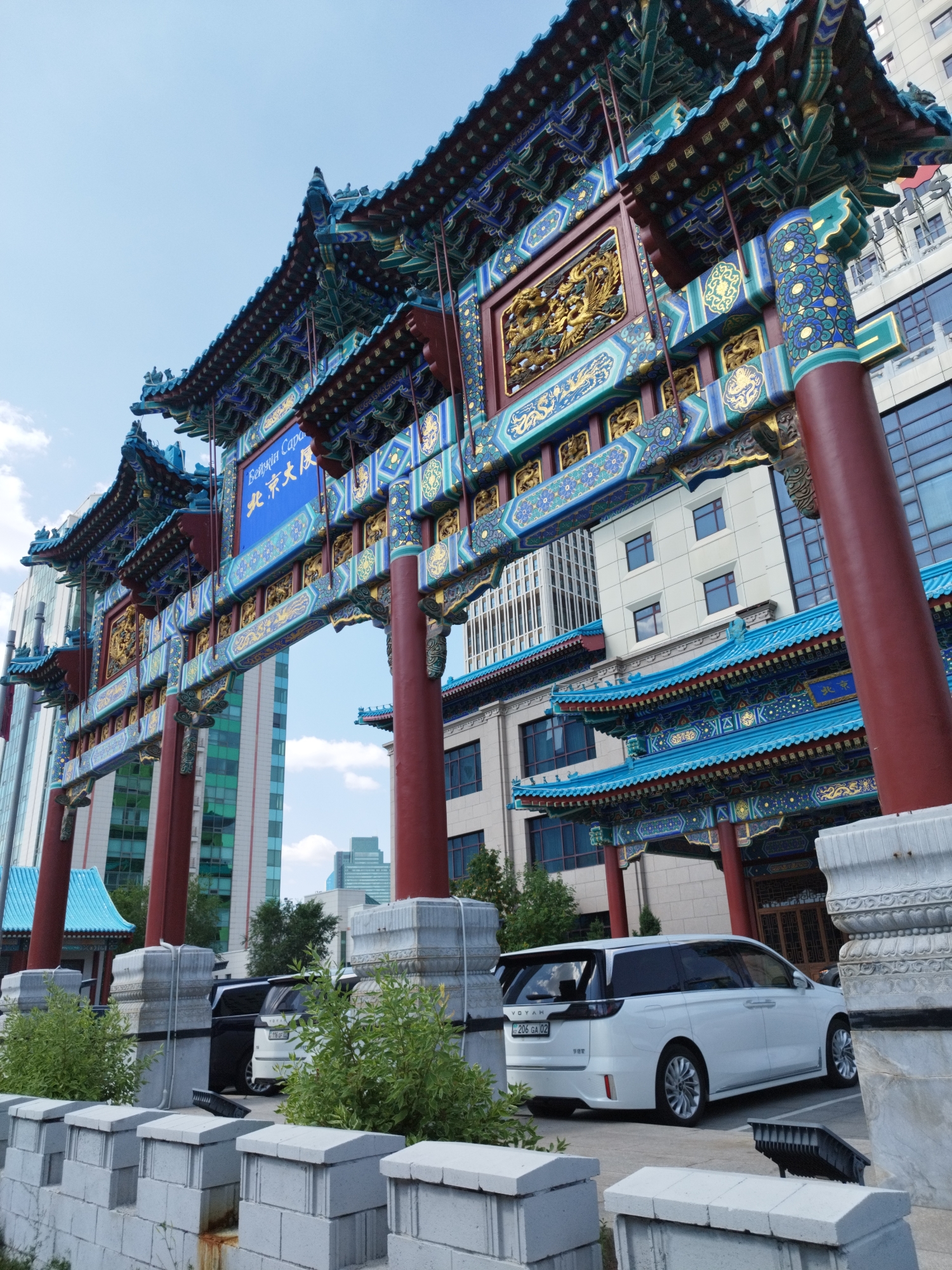 The image shows a detailed view of the "Beijing Capital" gate, a traditional Chinese archway located in Astana, Kazakhstan. The gate is elaborately decorated with blue, green, and red painted wood, featuring intricate carvings and gold accents. Behind the archway, modern buildings with glass facades are visible, creating a contrast between traditional and contemporary architecture. In the foreground, a white minivan with a license plate that appears to be in Cyrillic script and a black SUV are parked. Green bushes and a low stone wall with decorative elements are also present in the foreground, framing the scene. The sky is blue with scattered clouds. The overall impression is of an architectural landmark within a city setting.
