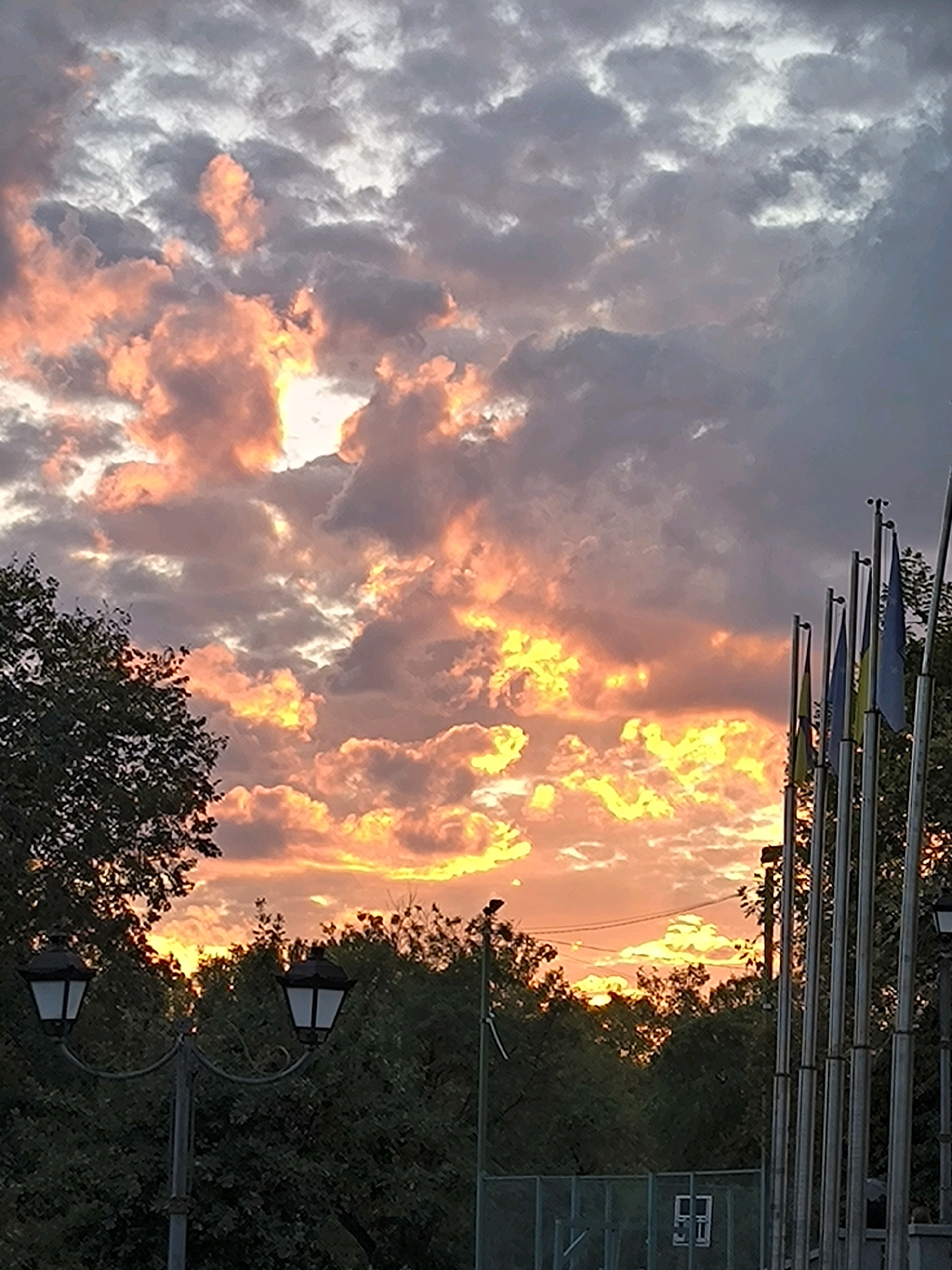 A dramatic sunset casts vibrant hues of orange and yellow across a cloudy sky. The sun is partially obscured by clouds, creating a brilliant, luminous effect. In the foreground, trees line the horizon, their dark silhouettes contrasting with the bright sky. To the left, a vintage-style lamppost with two lamps stands against the backdrop of the trees. On the right side of the image, a row of flagpoles with flags, some appearing to be blue and yellow, is visible. Further in the background, a chain-link fence and some structures can be discerned. The overall scene captures the beauty of a sunset in an urban park or public space, possibly in Mykolaiv, Ukraine, given the presence of Ukrainian flag colors.