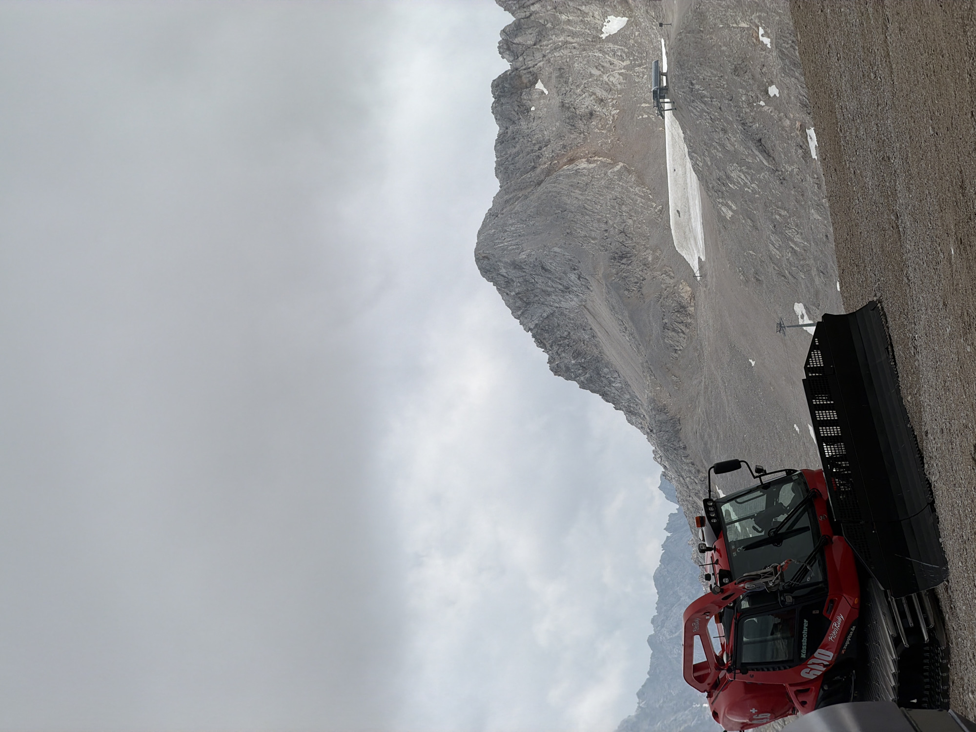 The image depicts a red snowcat vehicle positioned on a rocky, barren mountain slope. The vehicle, a Kassbohrer PistenBully 600, has "Zugspitze" branding visible, indicating its location at Zugspitze, Germany's highest peak, which is accessible from Garmisch-Partenkirchen. The sky is overcast with clouds. In the background, rugged, rocky mountains rise up, with patches of snow visible on their slopes and a ski lift station on a ridge. The ground is covered in gravel and loose stones. No people are visible in the frame. The scene is one of a high-altitude mountain environment, likely during the summer or shoulder season when snow grooming vehicles might be used for maintenance or preparing for early snow.