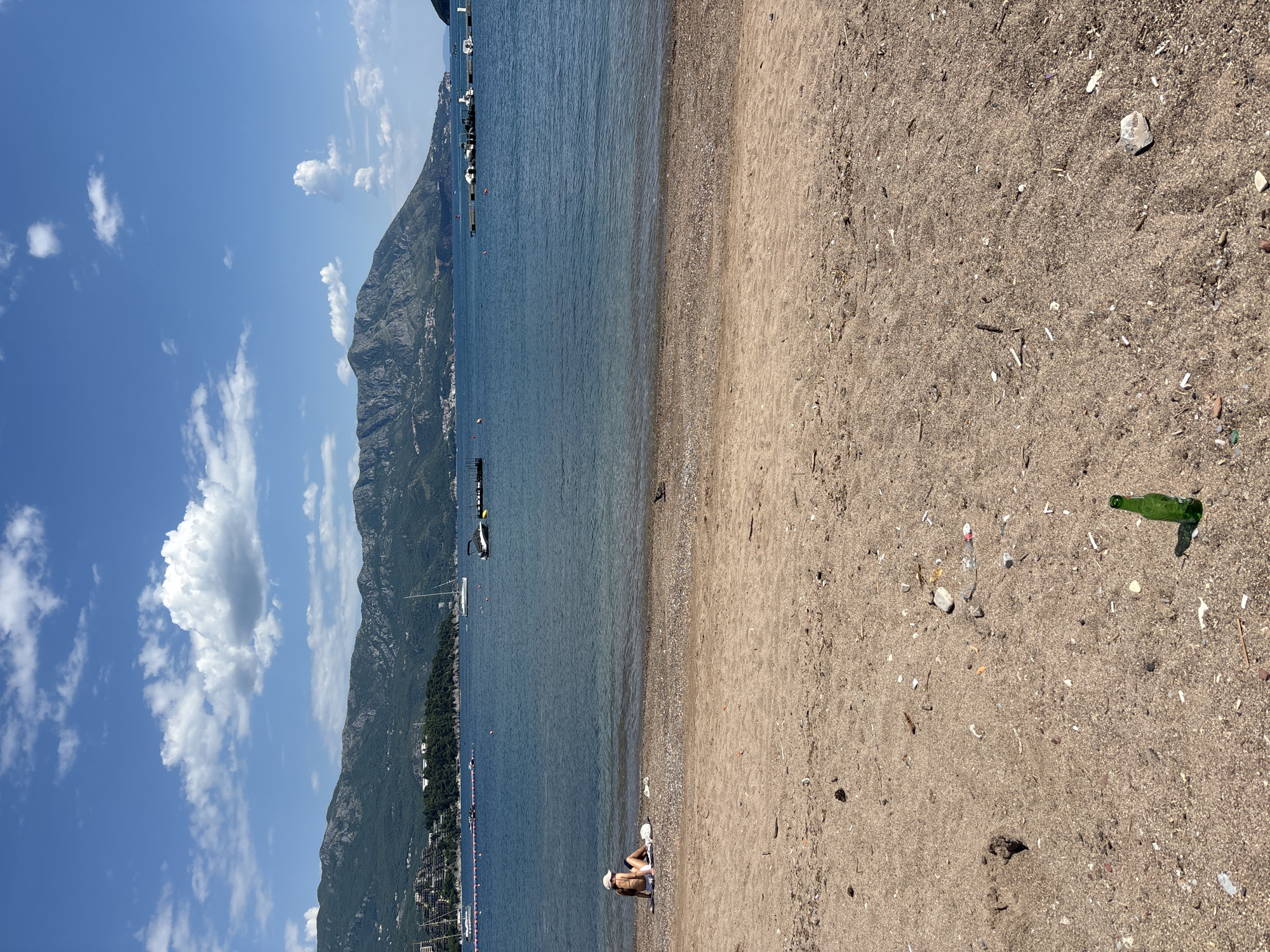 The image depicts a beach scene in Budva, Montenegro. The beach is sandy with visible litter scattered around, including a green glass bottle, small pieces of plastic, and shells. A person is sitting at the water's edge. In the background is the sea with boats. Beyond the sea are mountains. The sky is blue with some scattered clouds.