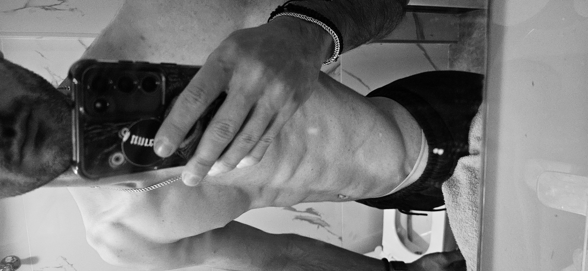 A black and white, low-angle reflection shows a man in a bathroom taking a selfie with a smartphone. The phone has a "RULE" pop-socket. The man wears a bracelet on his wrist. The background includes a sink, toilet, towel, and tiled walls. The location in Plovdiv, Bulgaria is unknown.