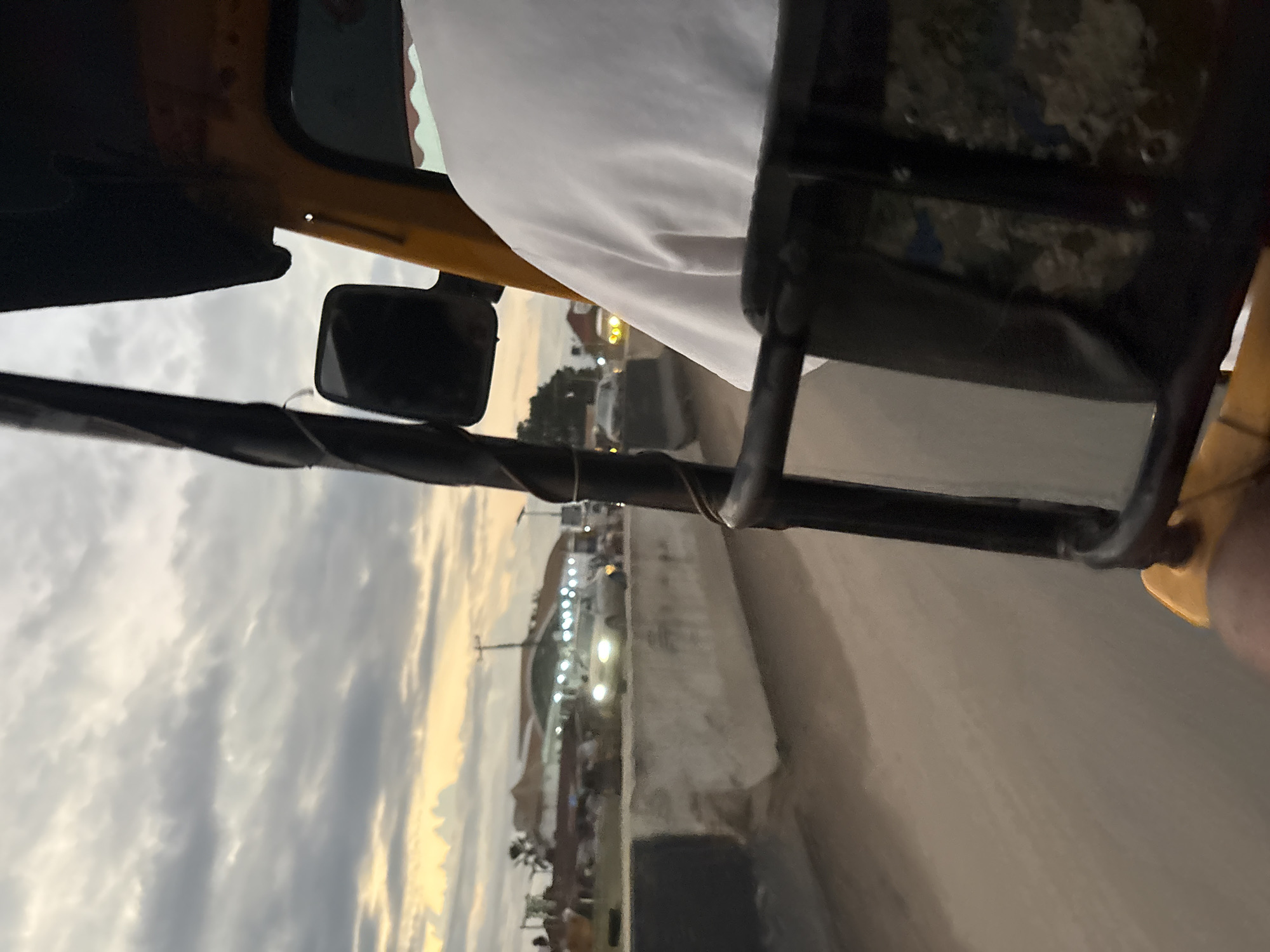 This is a point-of-view shot from the perspective of someone riding in an open-air vehicle, likely a motorized rickshaw, in Onitsha, Nigeria. The time of day appears to be dusk or early evening, with a dramatic cloudy sky showing patches of yellow and orange light near the horizon, suggesting the sun is setting.
The vehicle is a bright yellow and has a black frame. A portion of the driver's white shirt is visible in the foreground, suggesting the passenger is seated behind the driver. The vehicle's black metal frame and a side mirror are prominent elements in the foreground.
The road ahead is unpaved and dusty. On the right side of the road, there's a concrete barrier, partially covered with what appears to be black and white material. Beyond the barrier, several vehicles are visible, including a car with its headlights on, moving in the opposite direction. In the distance, buildings are lit up, indicating a populated area. Trees and streetlights are also visible. The overall impression is of a busy urban environment at the close of day, with the characteristic atmosphere of a Nigerian city.