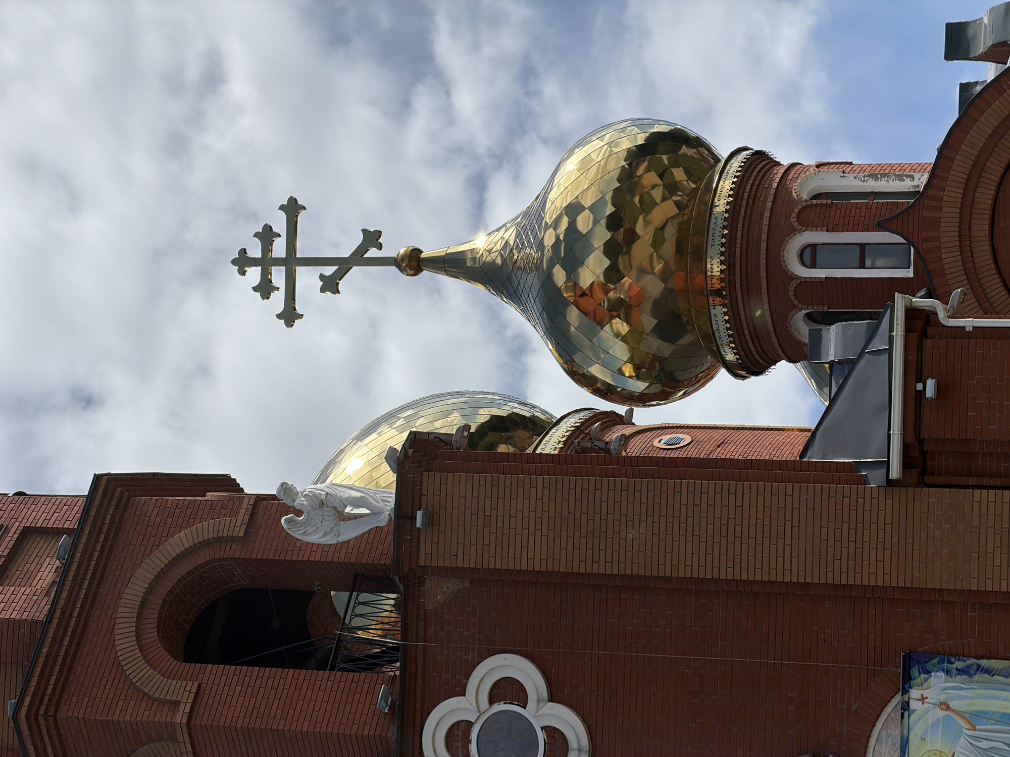 A low-angle shot presents the exterior of the Orthodox Church of St. George the Victorious in Chaikovsky, Russia, from a slightly rotated perspective. The church's architecture is predominantly brick, with two golden onion domes dominating the upper portion of the frame. The larger, more prominent dome is topped with an Orthodox cross. Below, an angel statue adorns the brick facade. Patches of sky and clouds are visible in the background.